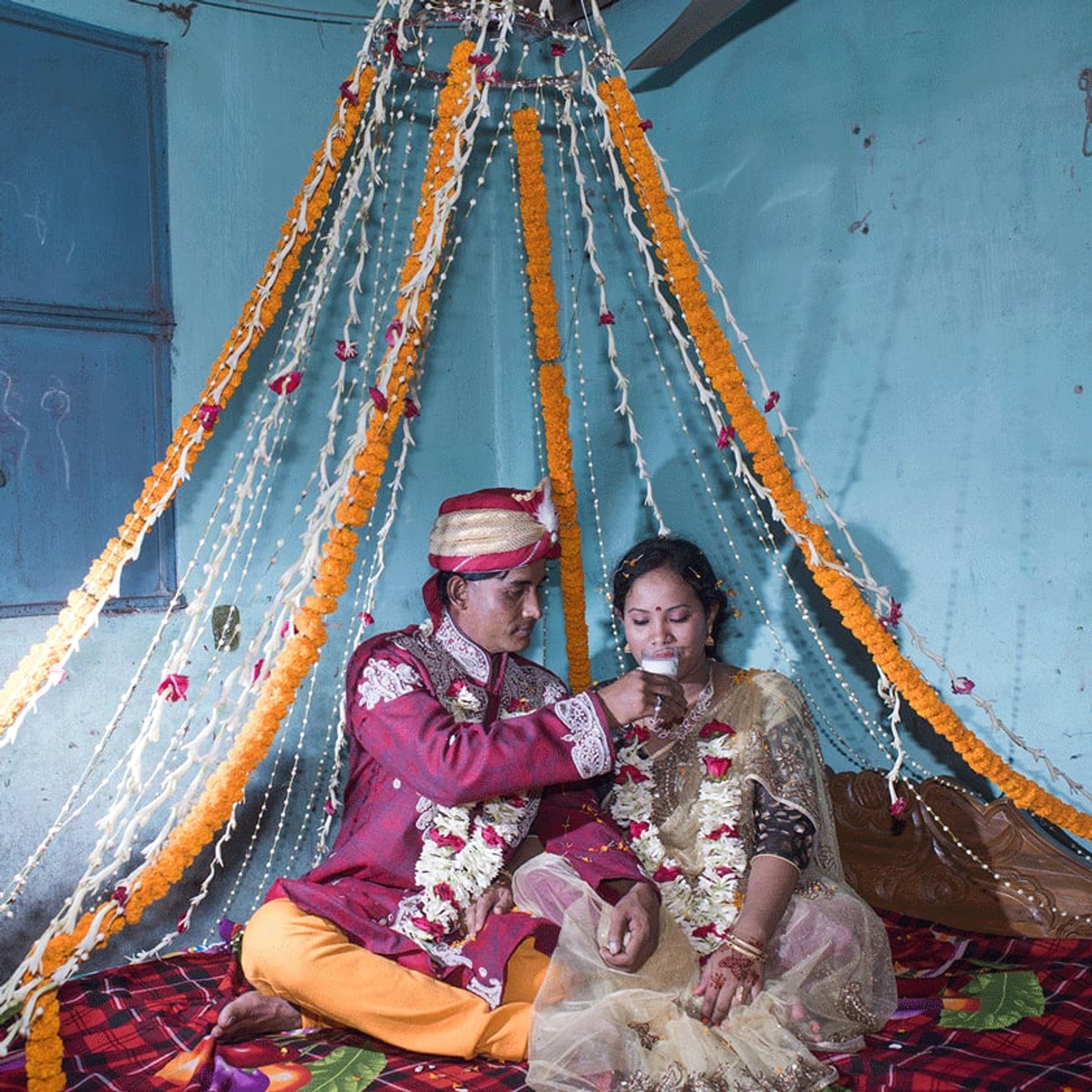 Two adults in wedding attire sit together beneath garlands on a decorated bed.
