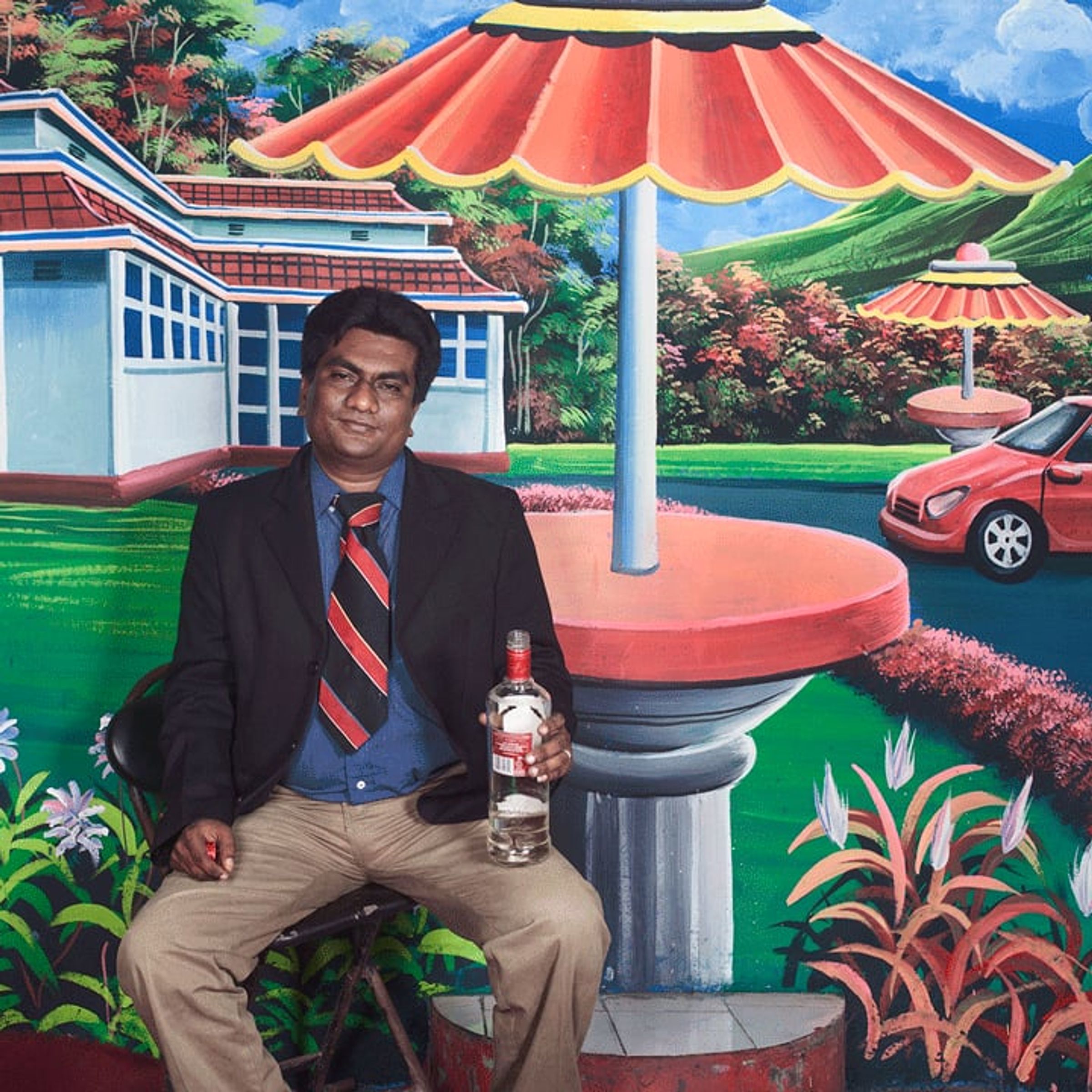 Man in a suit holds a bottle while seated before a painted pavilion and garden.