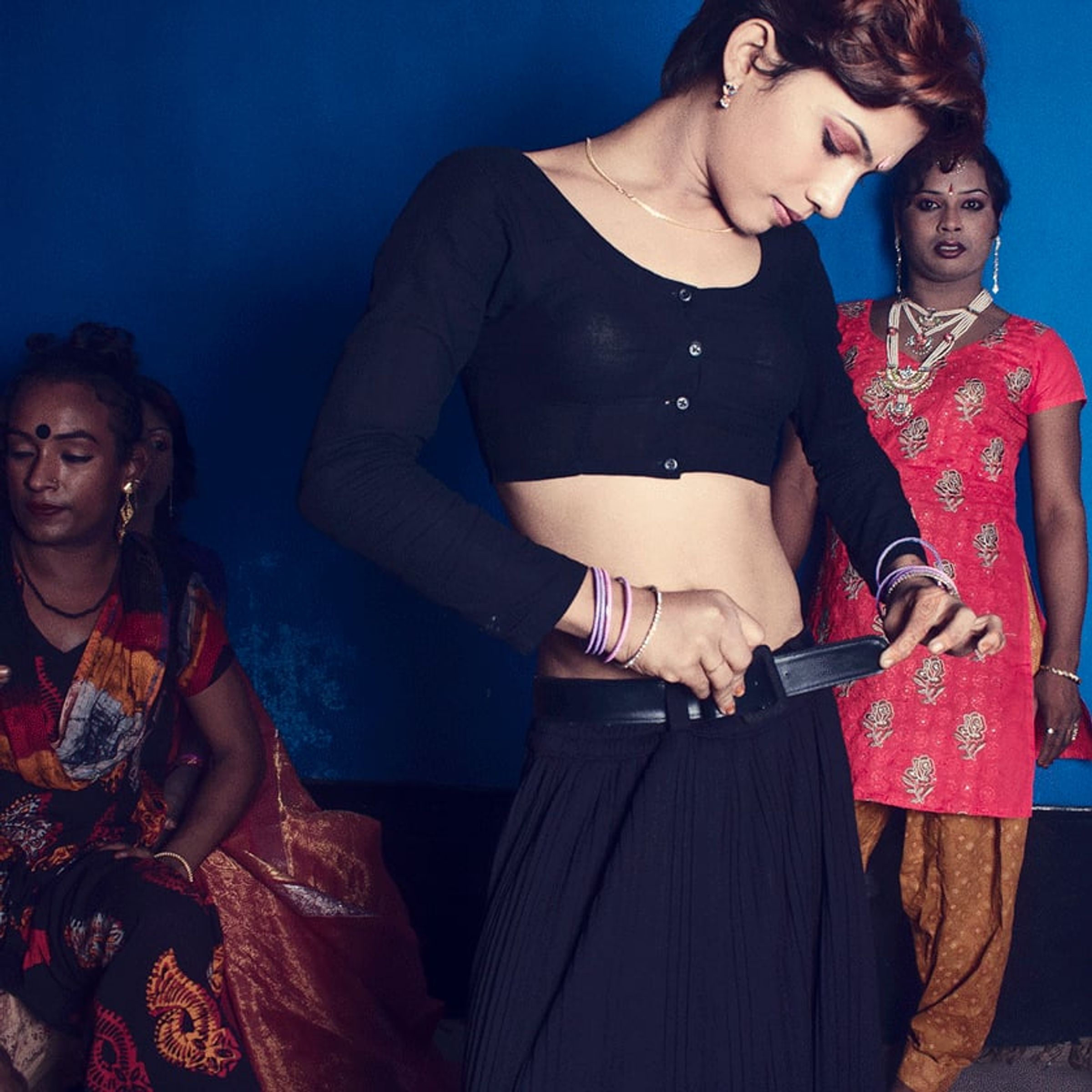 Person in black looks downward while brightly dressed figures stand behind against a blue backdrop.