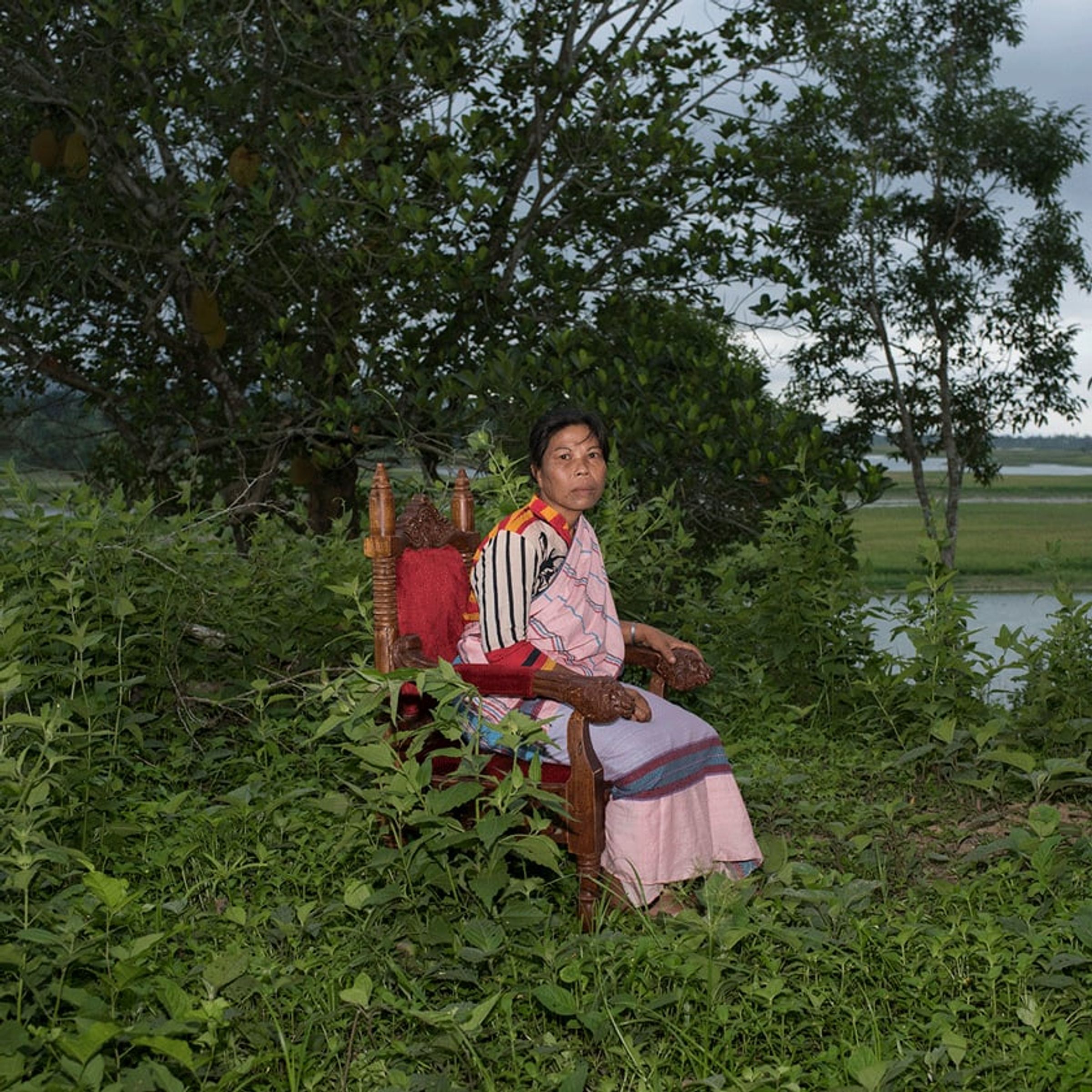 Seated person in striped clothing rests on an ornate red chair surrounded by vegetation.