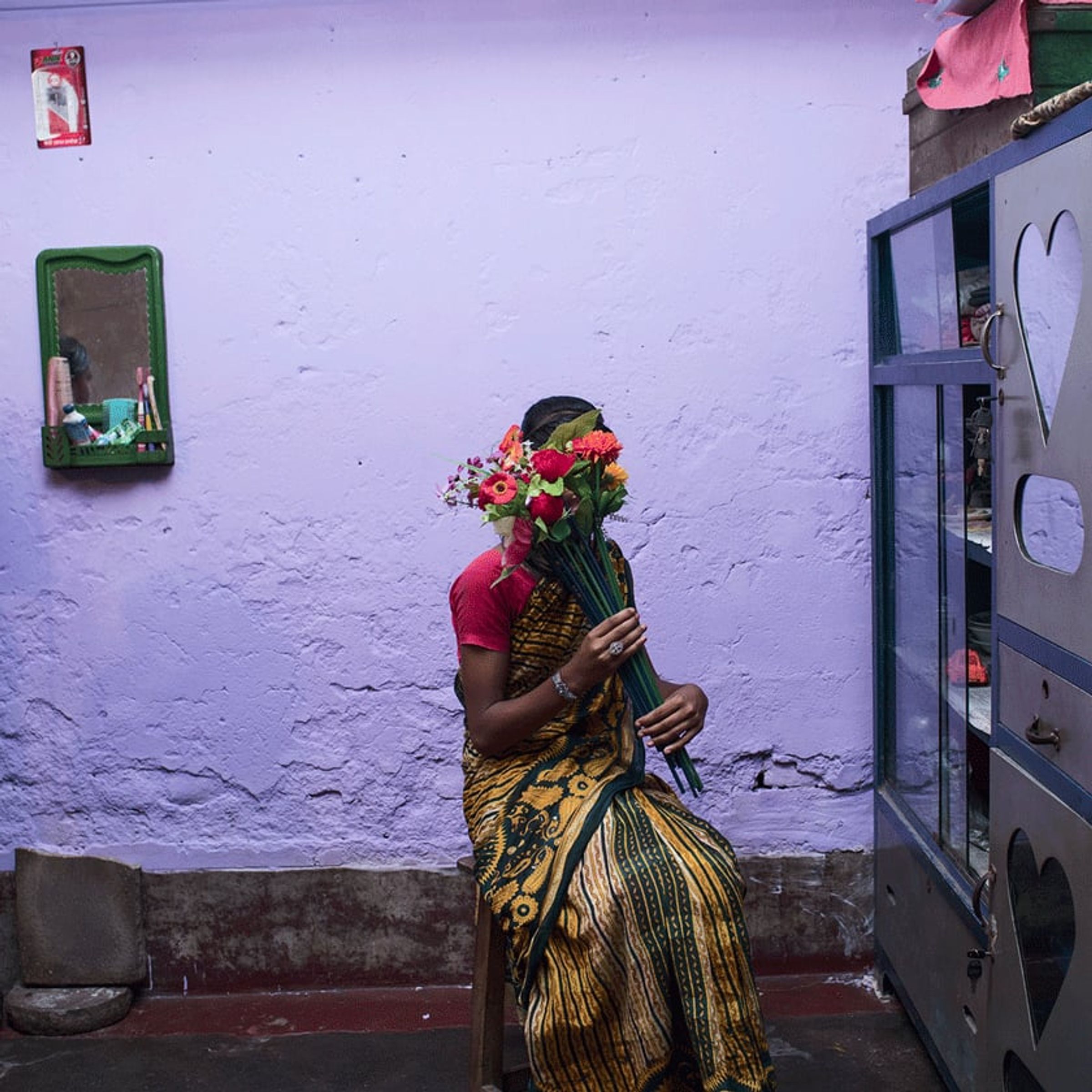 Person in a patterned sari carries a bouquet through a purple-painted courtyard.
