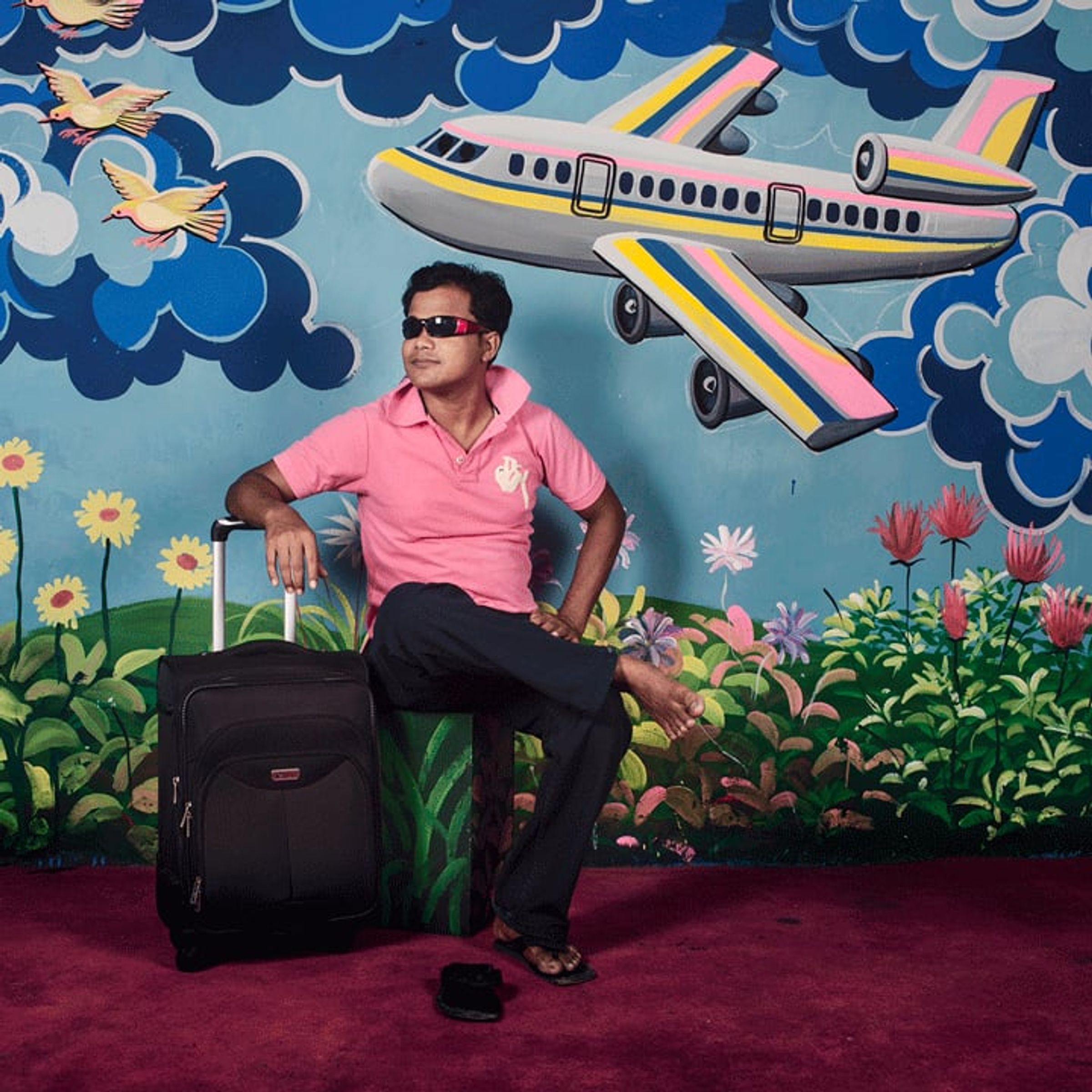 Seated adult in sunglasses poses beneath a painted airplane and storm-cloud backdrop.