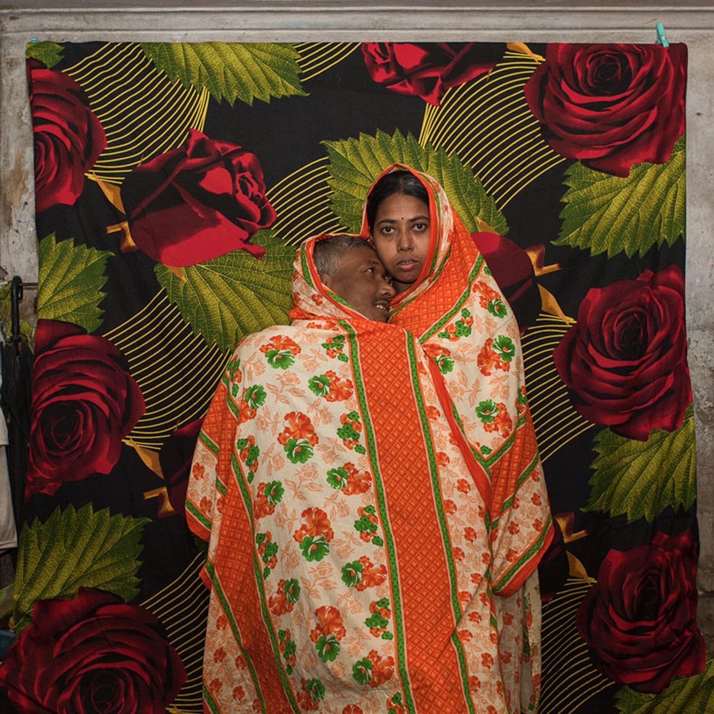 Adult wrapped in floral-patterned cloth stands against a backdrop of oversized red roses.