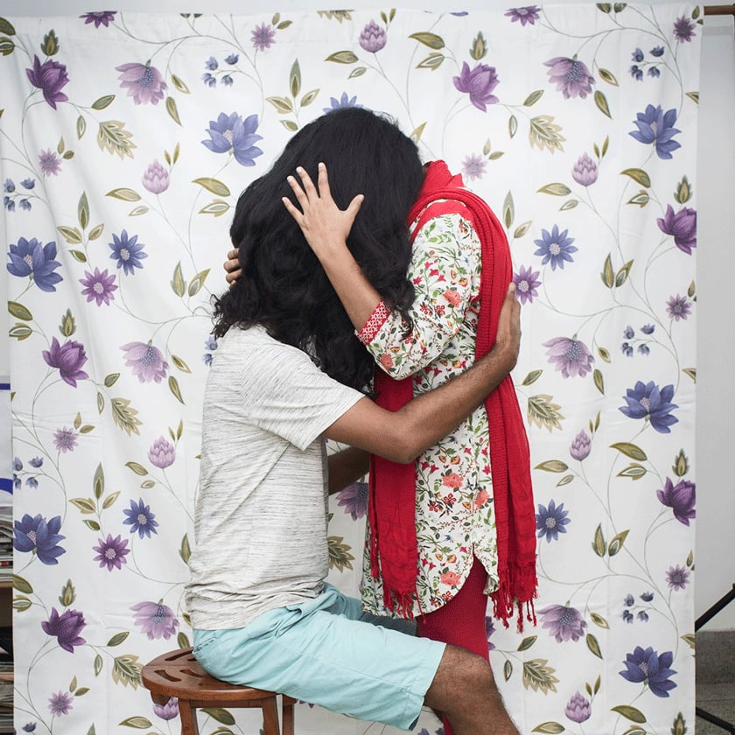 Kneeling couple embraces and kisses before a pale floral fabric backdrop.