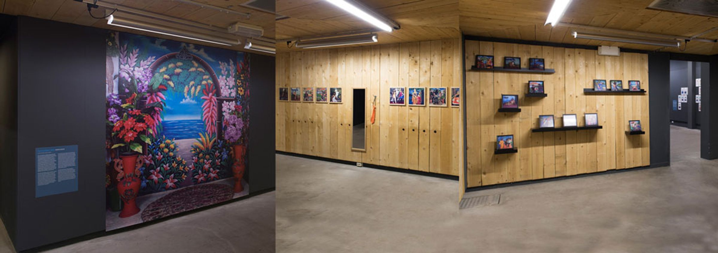 Gallery installation with a painted floral backdrop, framed works, and objects displayed along wooden walls.