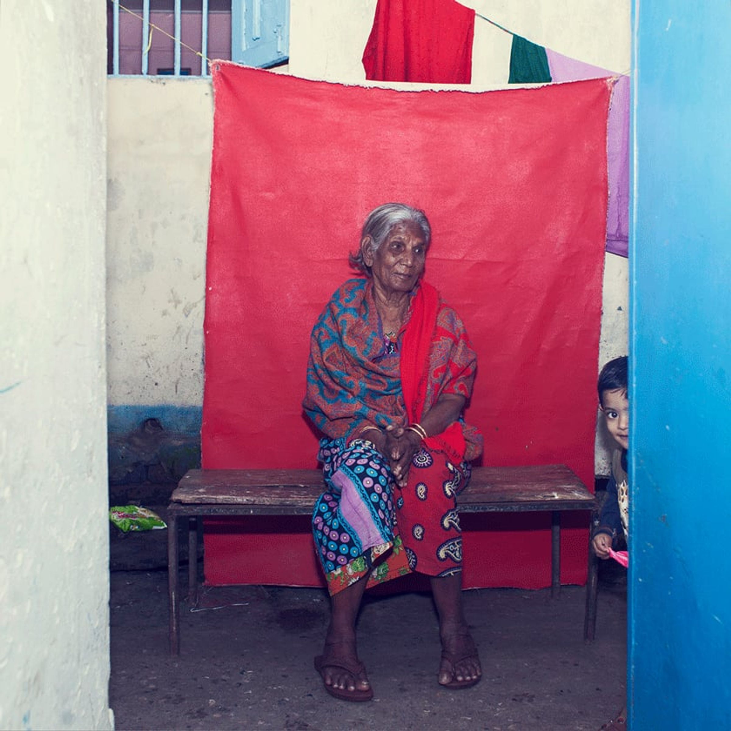 Elderly person sits on a wooden bench before a red backdrop.