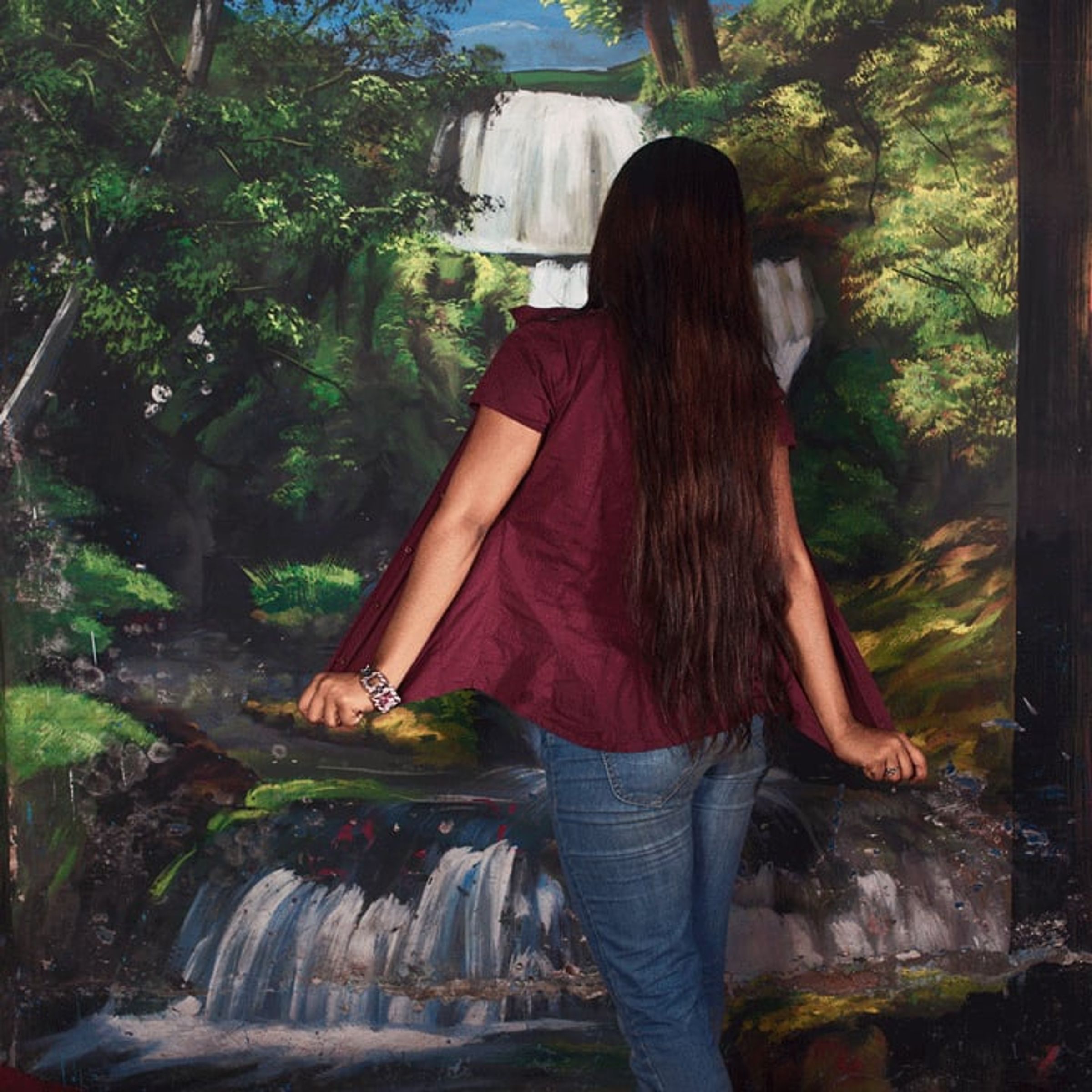 Long-haired person faces away toward a painted waterfall and wooded landscape.