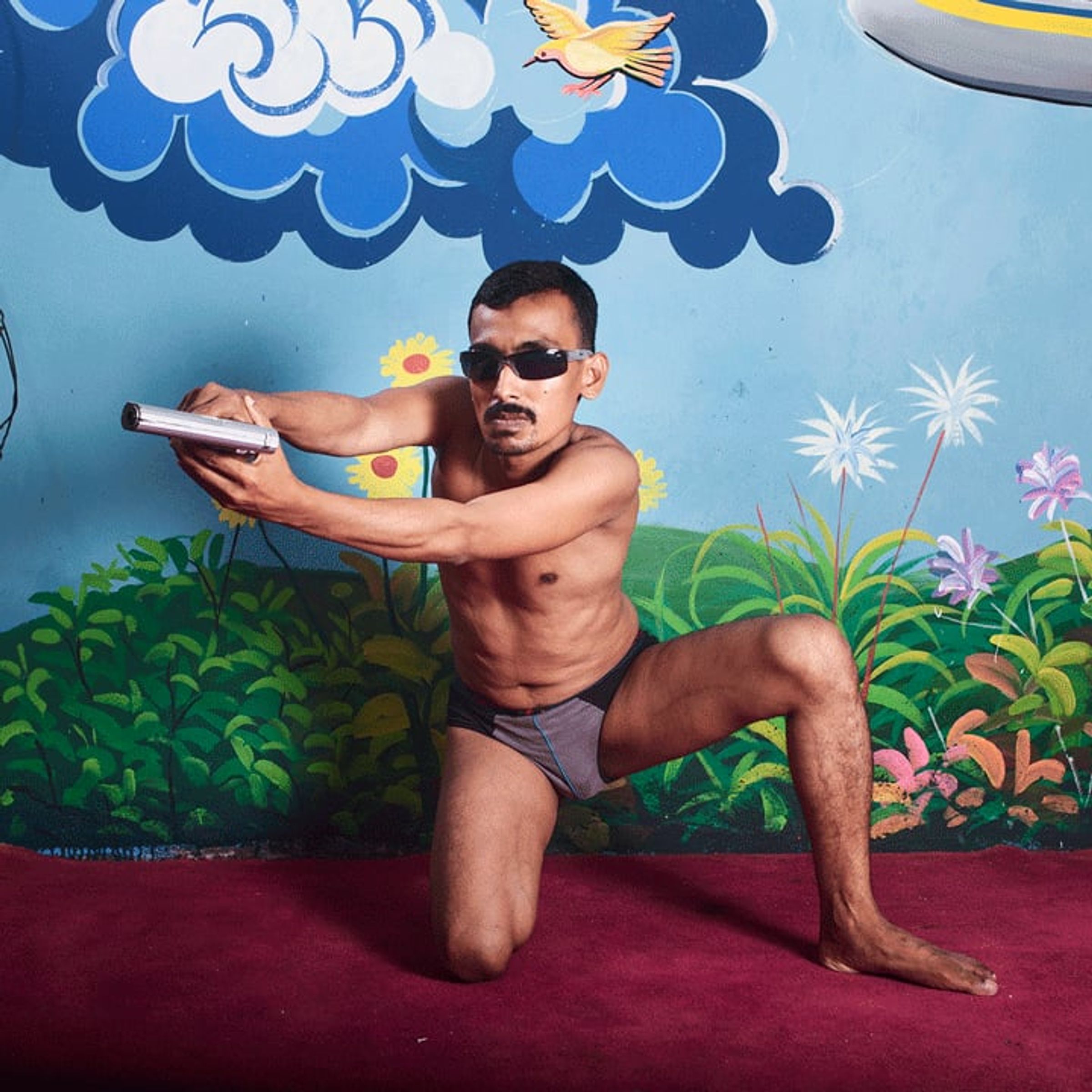 Shirtless man kneels with a toy gun before a painted airplane and sky.