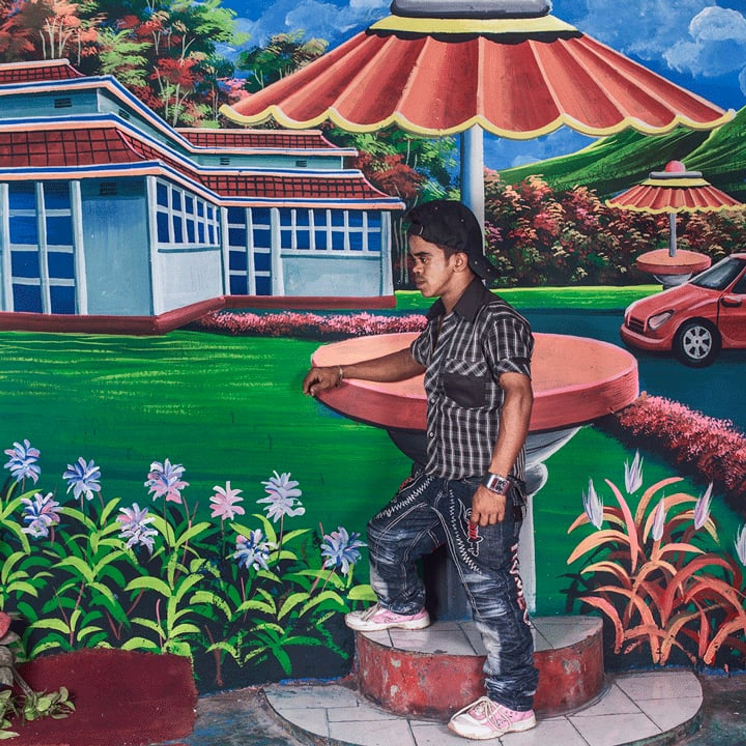 Young man stands beside a painted fountain, pavilion, and red car.