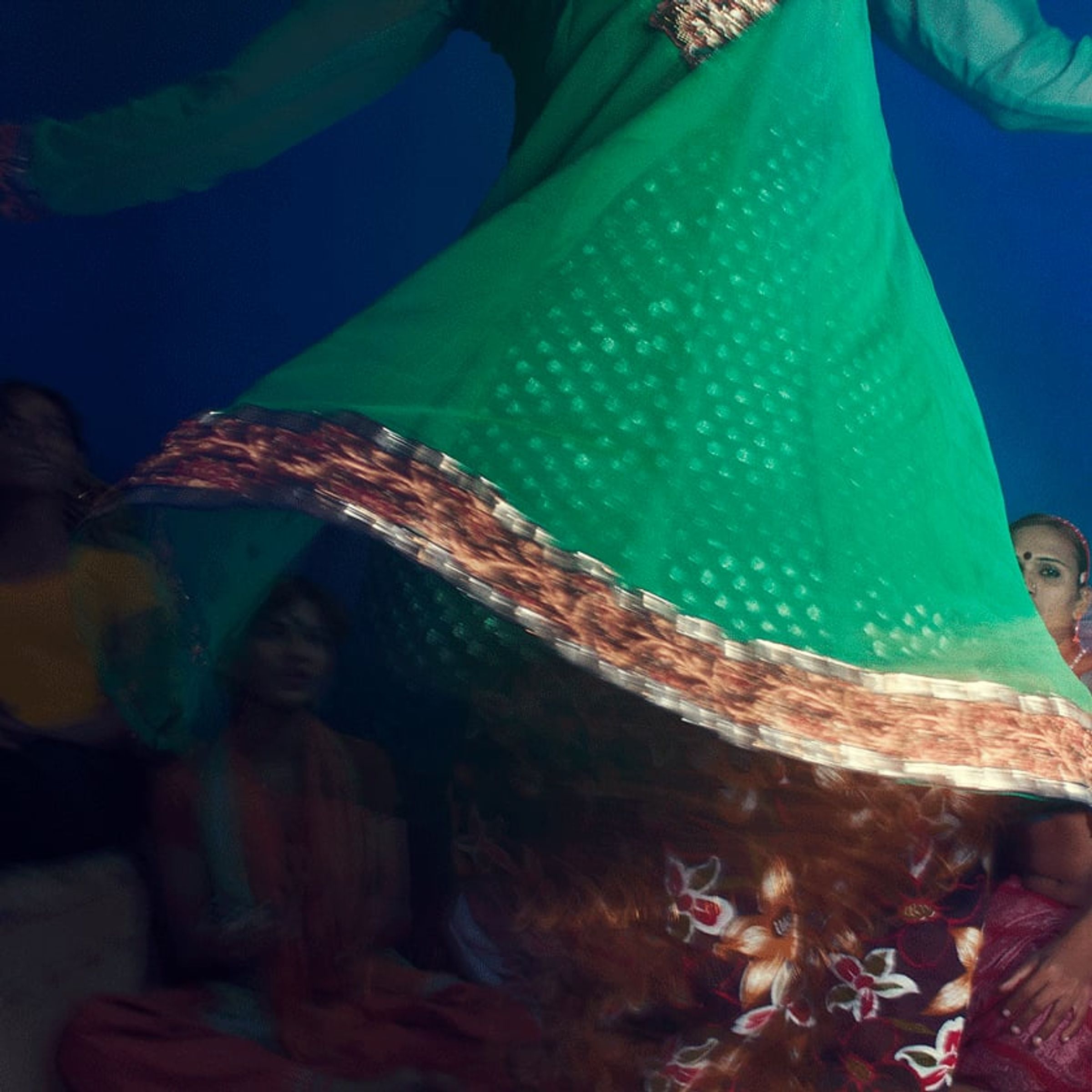Green patterned fabric swirls close to the camera, partially obscuring people seated against blue.