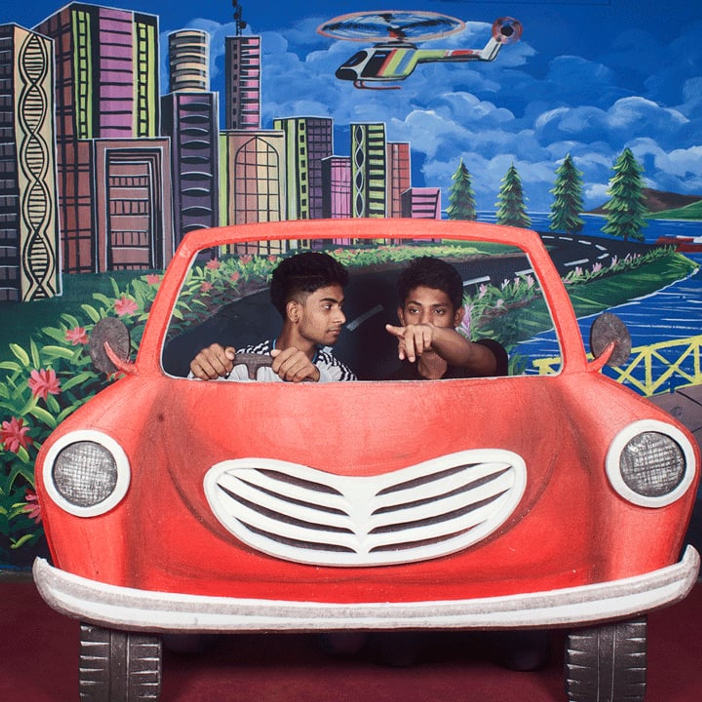 Two young men pose inside a painted red car against a city-and-sky backdrop.