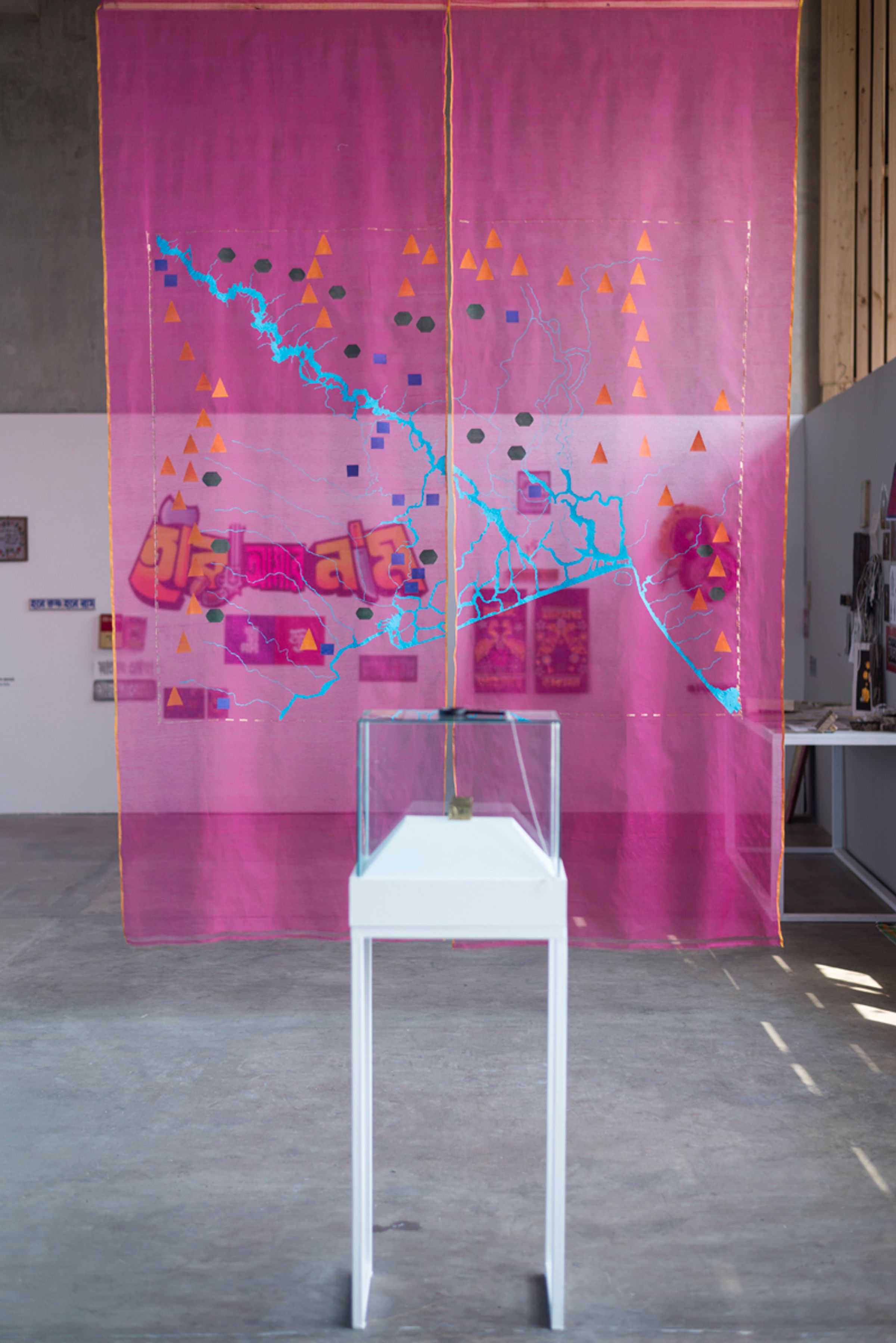 Translucent pink textile map hangs behind a white pedestal in an exhibition space.