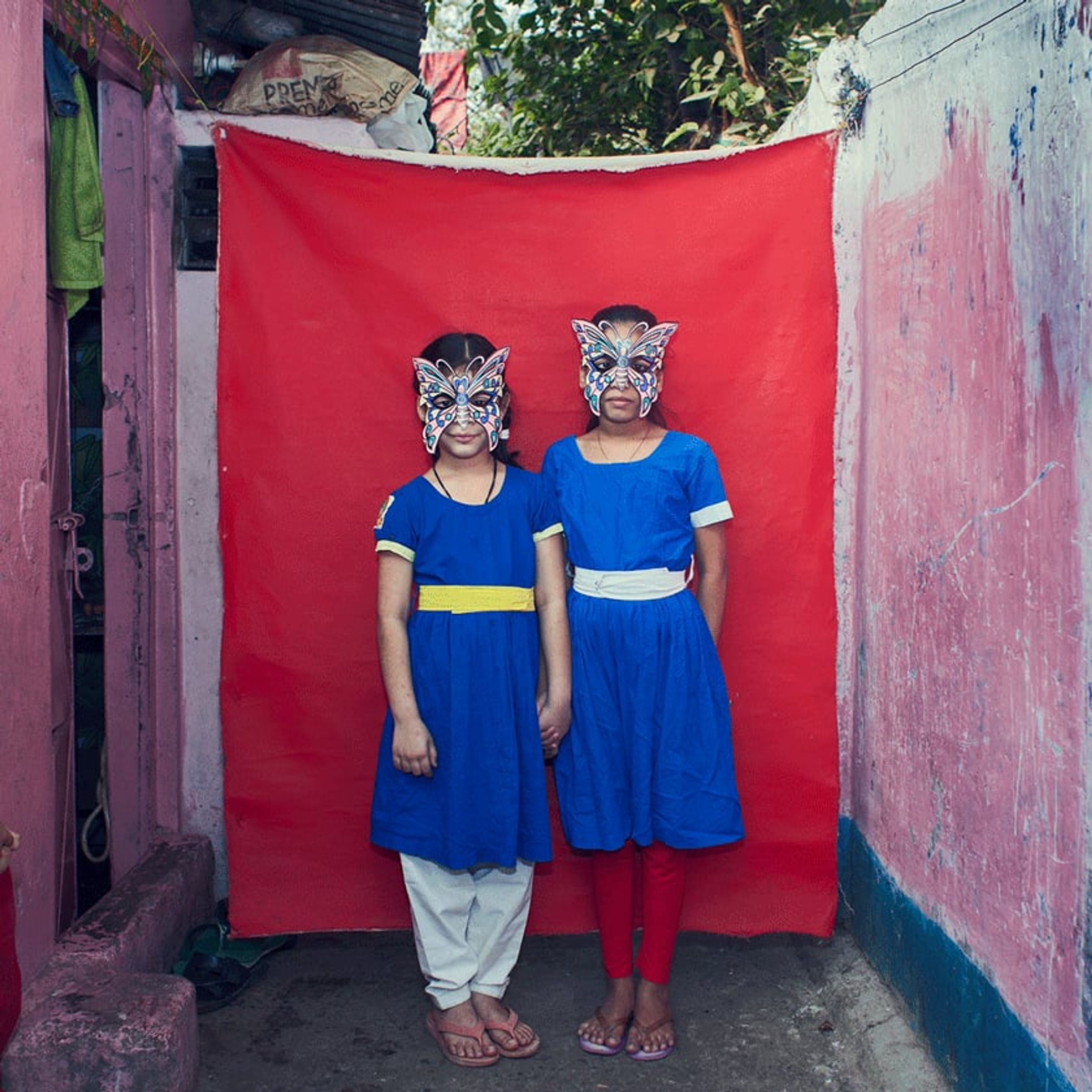 Two young people in matching blue uniforms wear painted animal masks before red cloth.