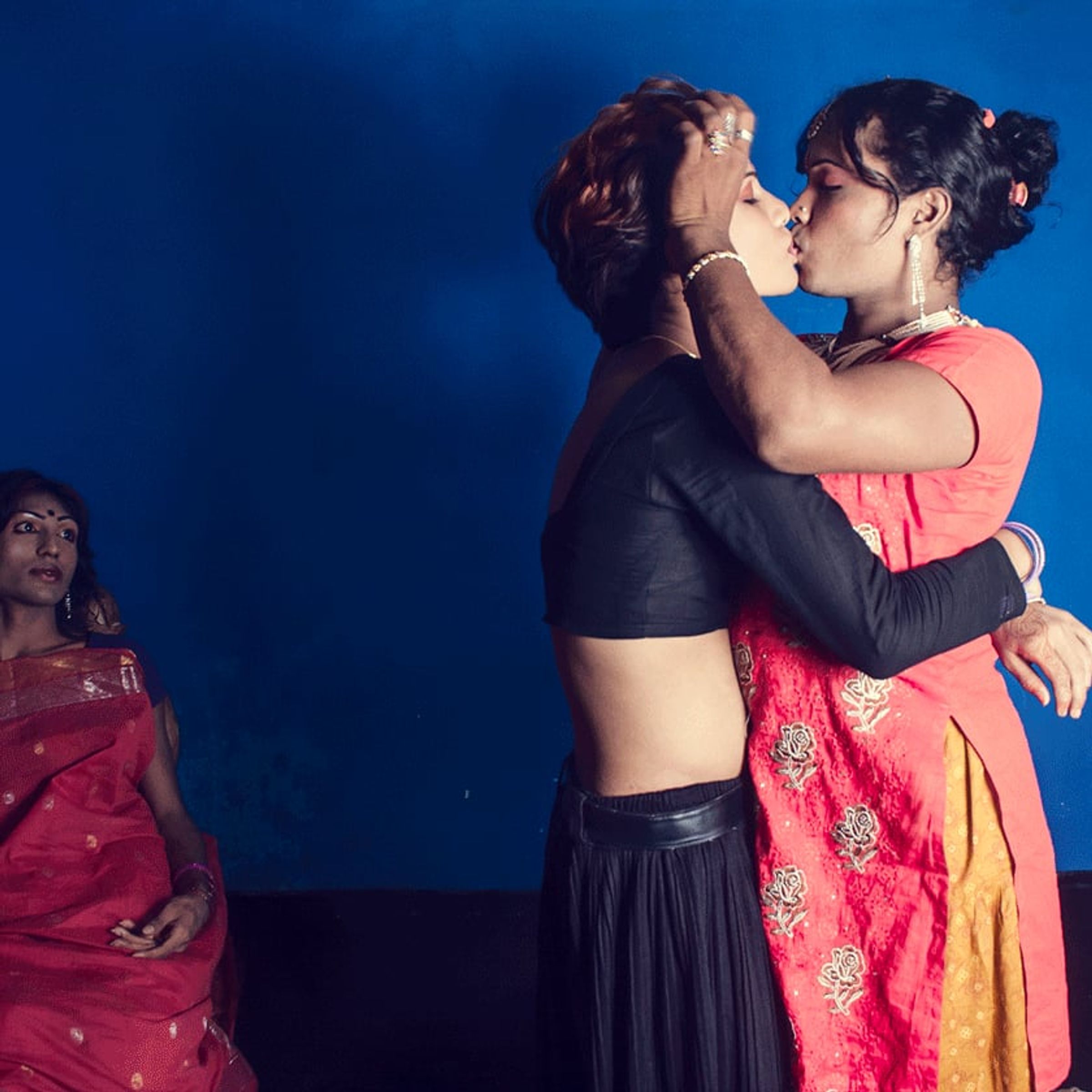 Two adults embrace and kiss before a blue backdrop as another person watches.