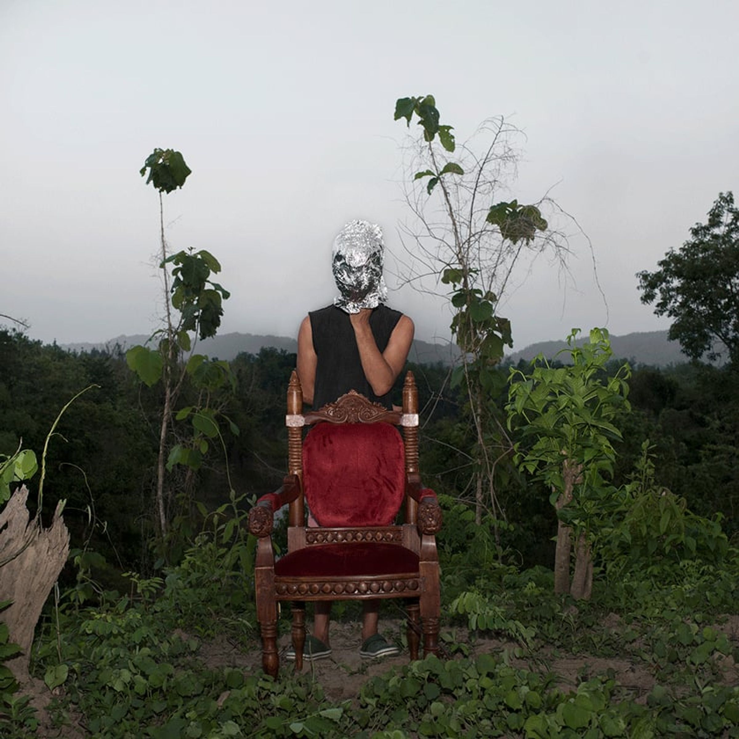 Seated figure faces away on a red chair, head wrapped in reflective silver material.