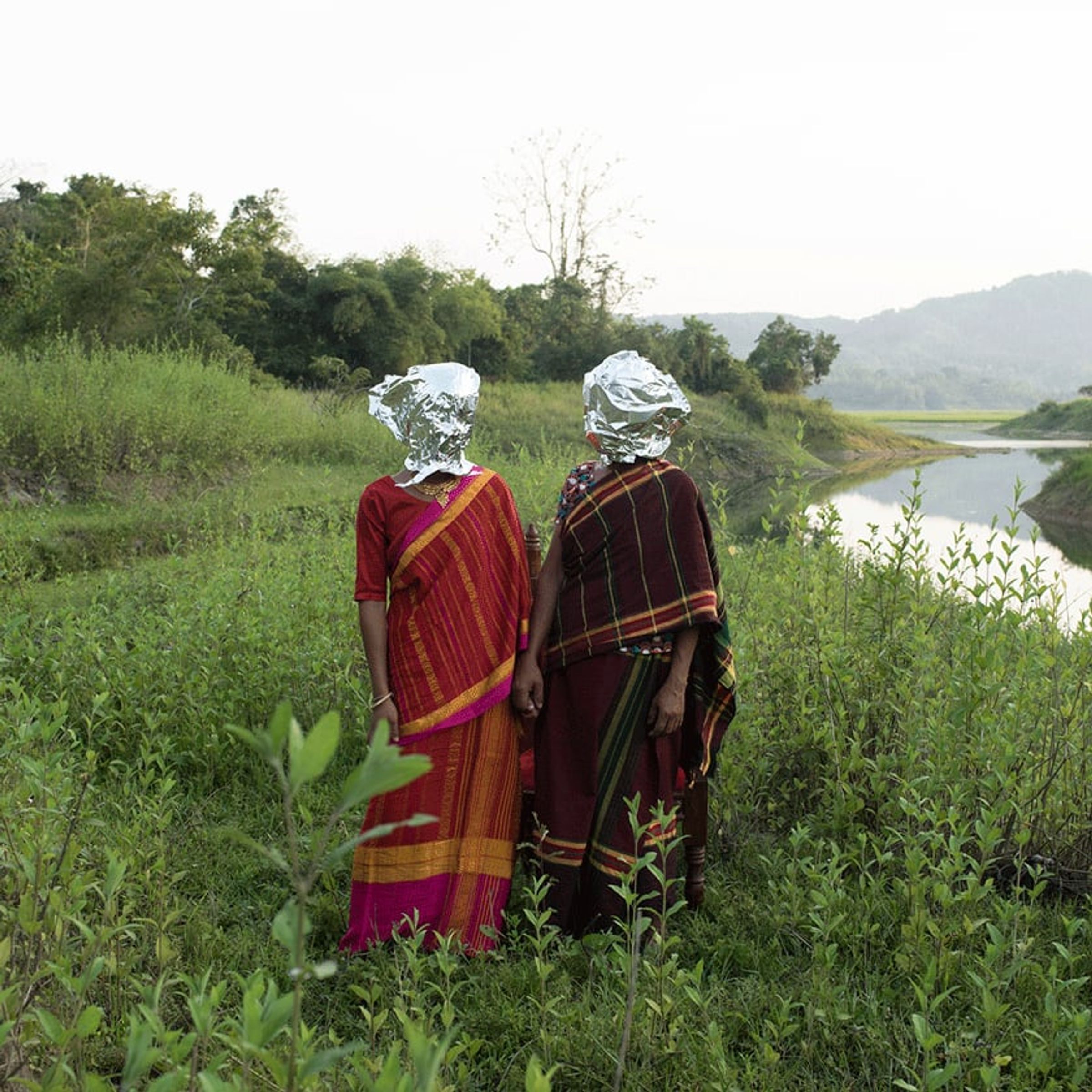 Two sari-clad figures with reflective silver head coverings face a river landscape.