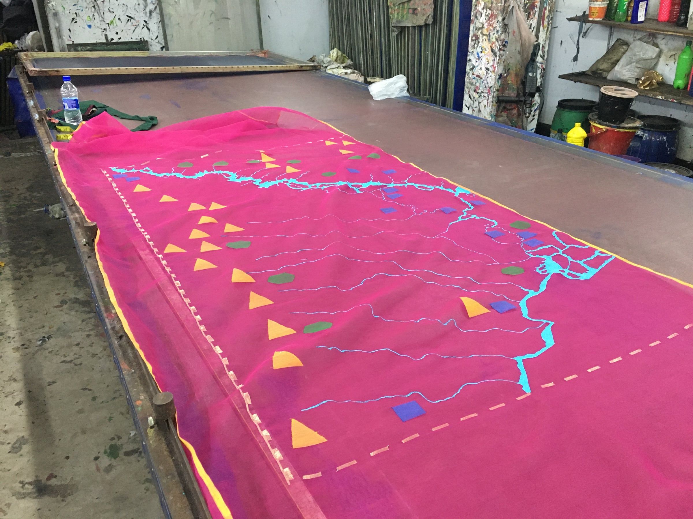 Bright pink Jamdani textile patterned with a blue river-like line and colorful geometric forms across a worktable.
