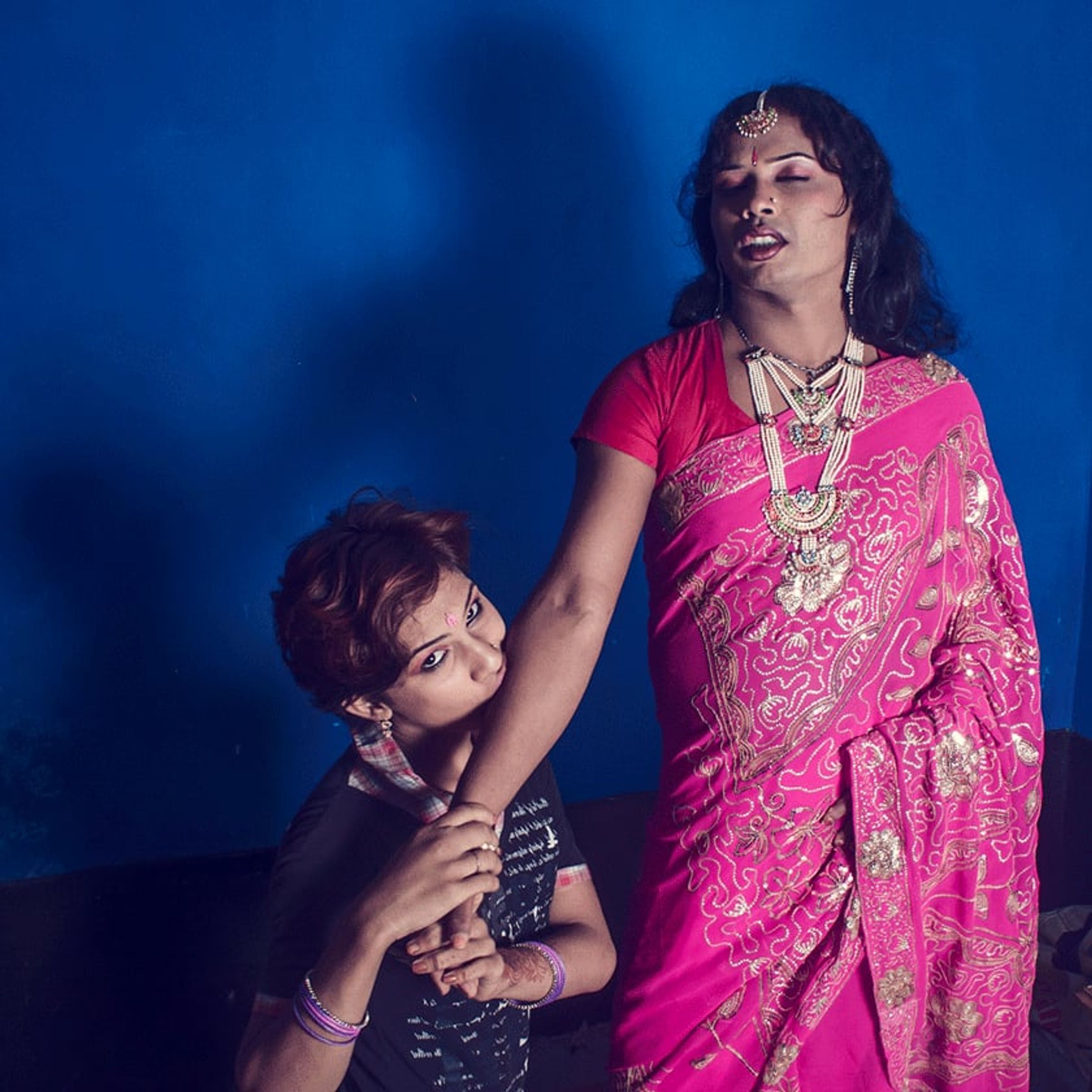 Seated person rests their head against a standing person in a pink sari.