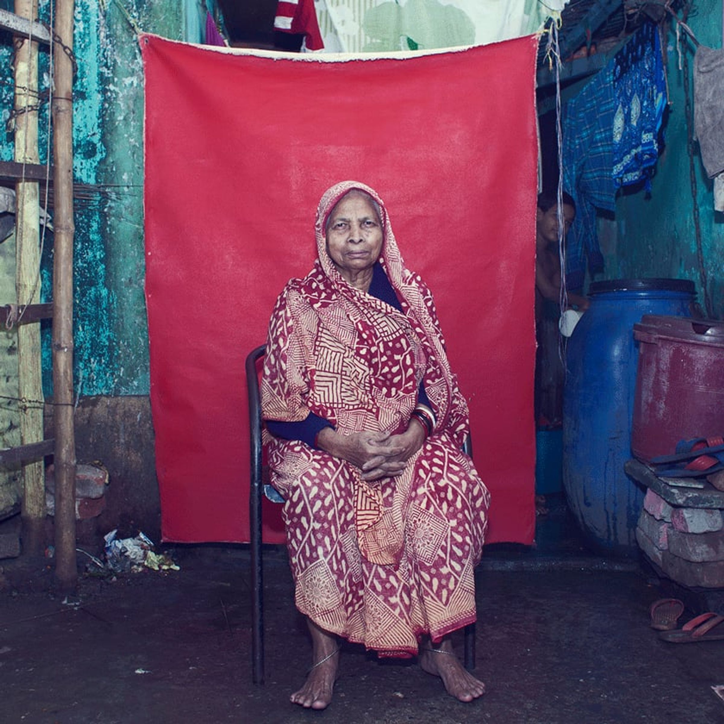 Elderly person in patterned red clothing sits squarely before a red fabric backdrop.