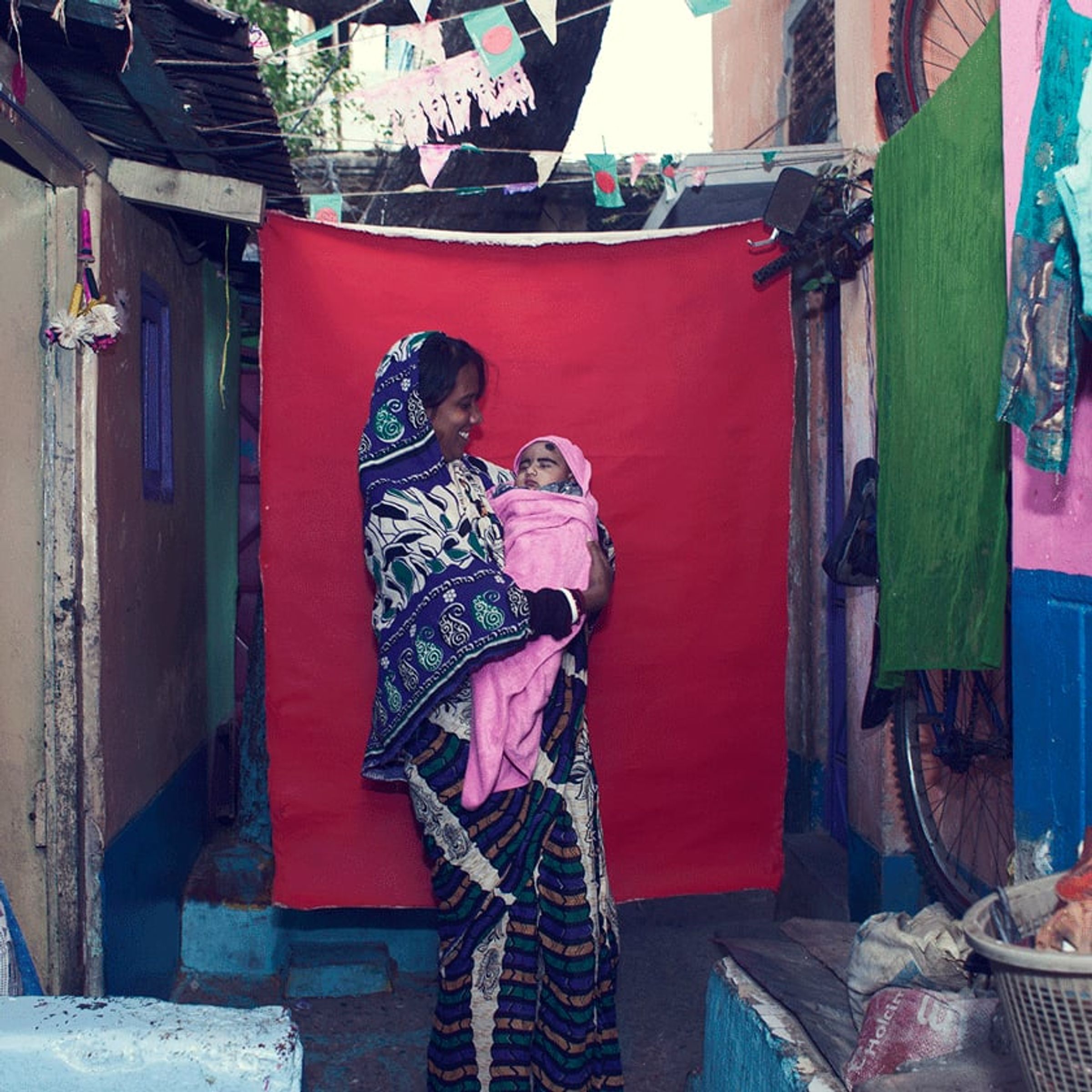 Adult holds a bundled infant in a narrow residential alley before red fabric.