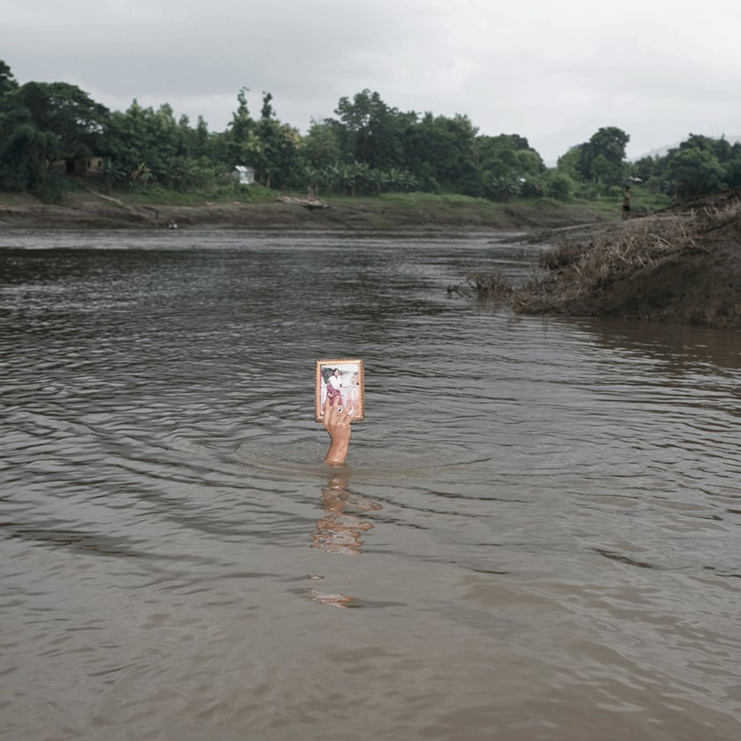 Person stands waist-deep in a river holding a framed portrait above the water.