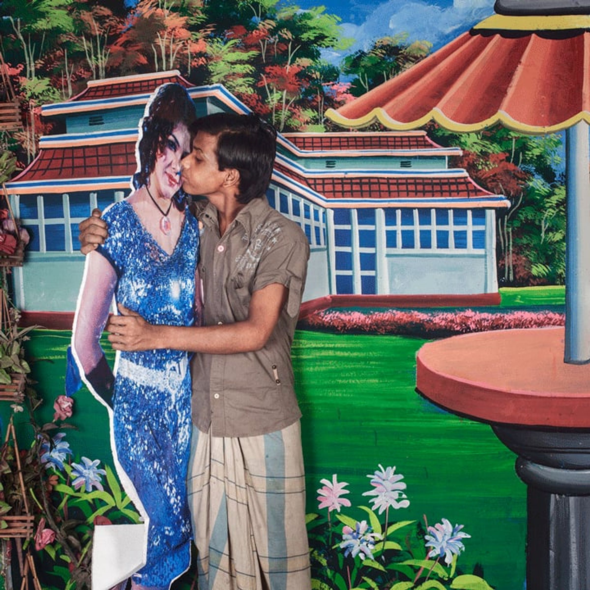 Couple embraces and kisses before a painted pavilion and garden backdrop.