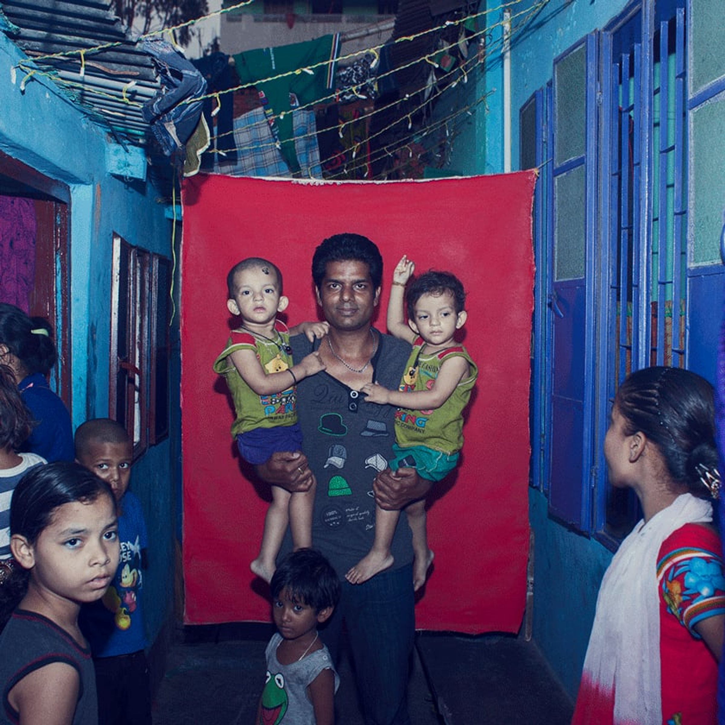 Adult holds two small children as neighbors gather around the red backdrop.