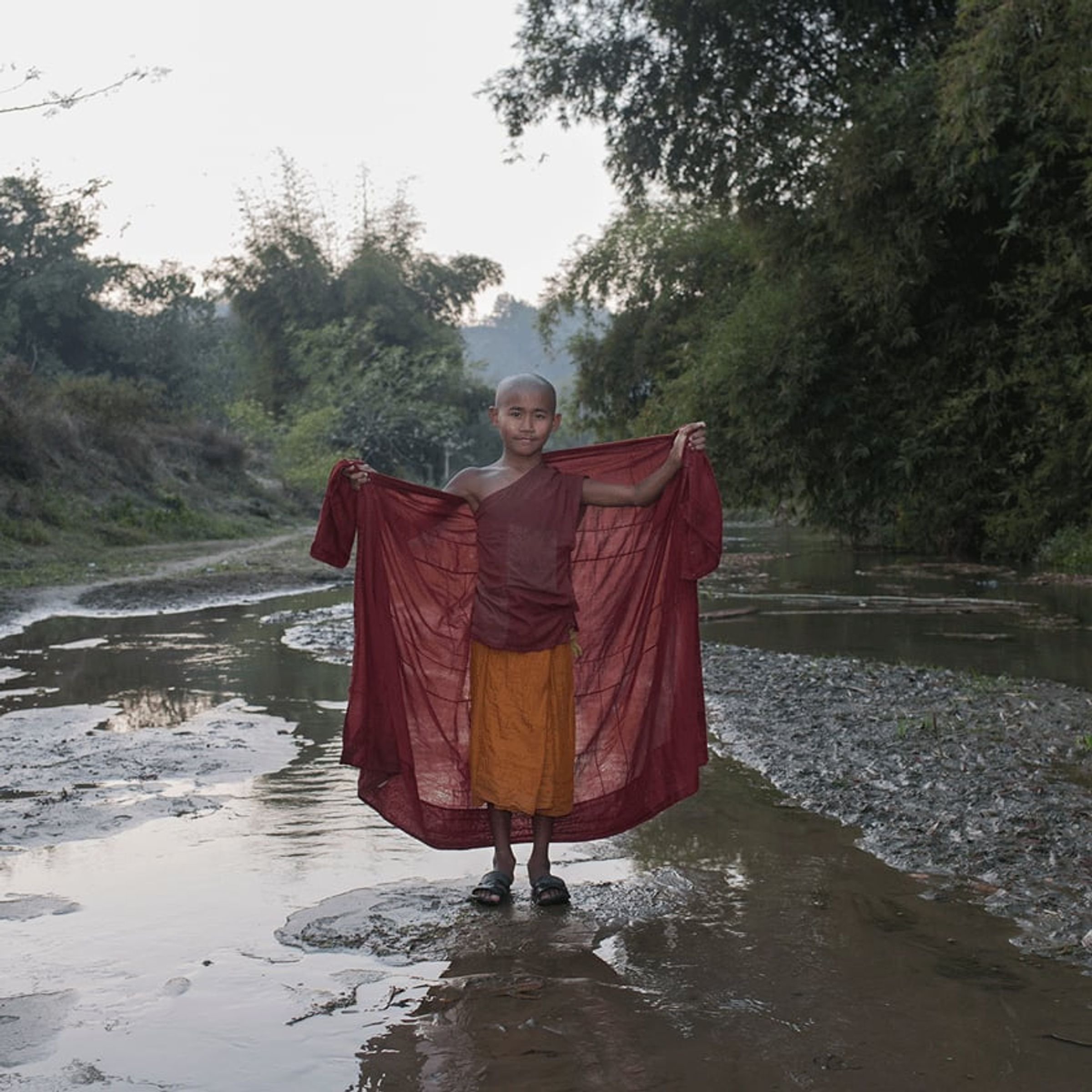 Shaved-head person stands in a stream holding a wide red cloth behind them.