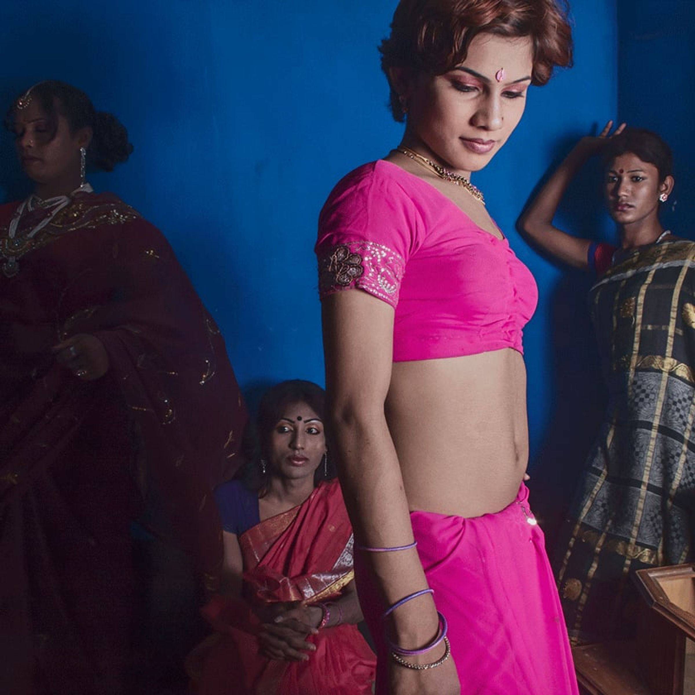 Person in a pink sari poses at center while other figures remain in shadow.