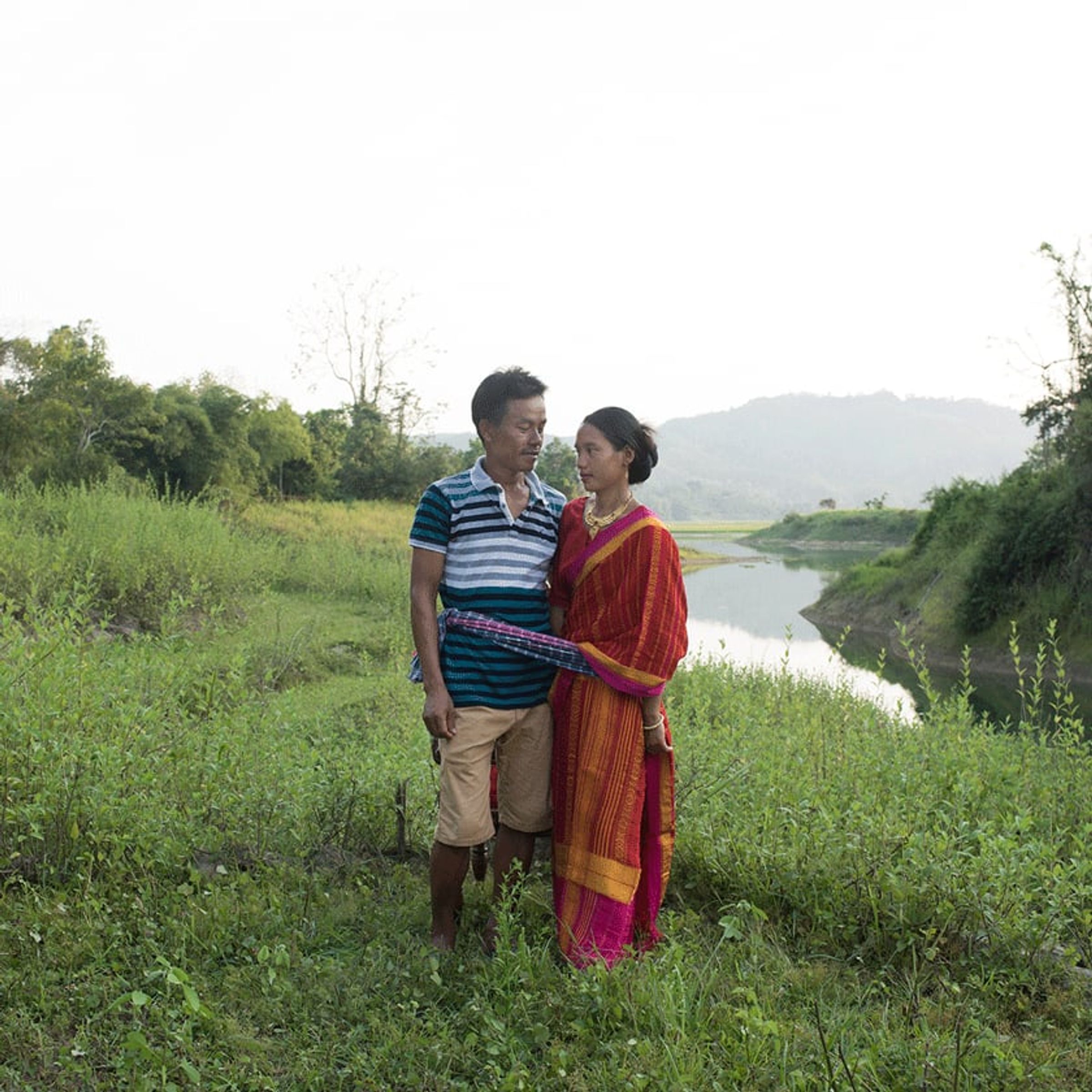 Two adults stand close together on a grassy bank beside a winding river.