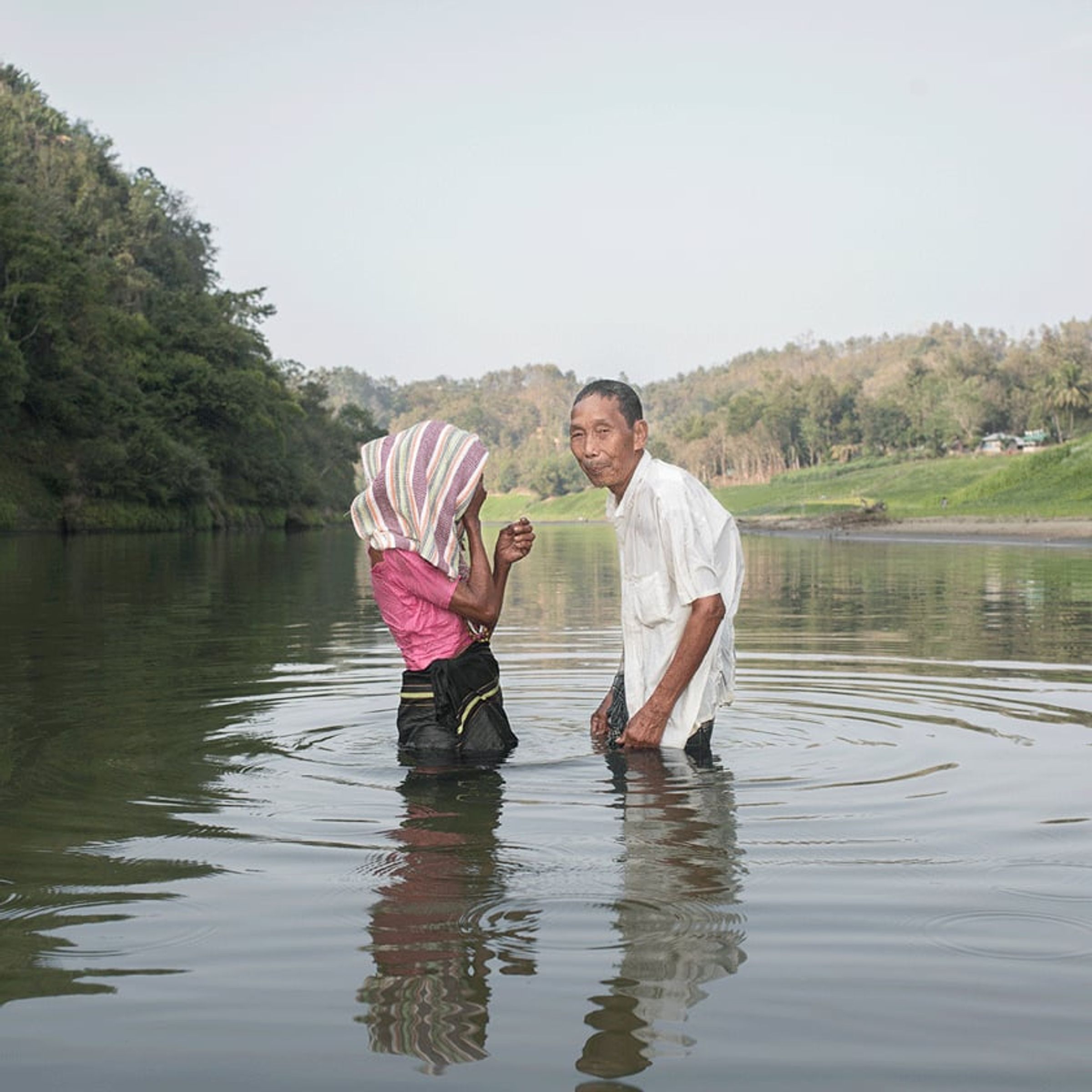 Two adults stand in shallow water, one lifting striped fabric around the other.