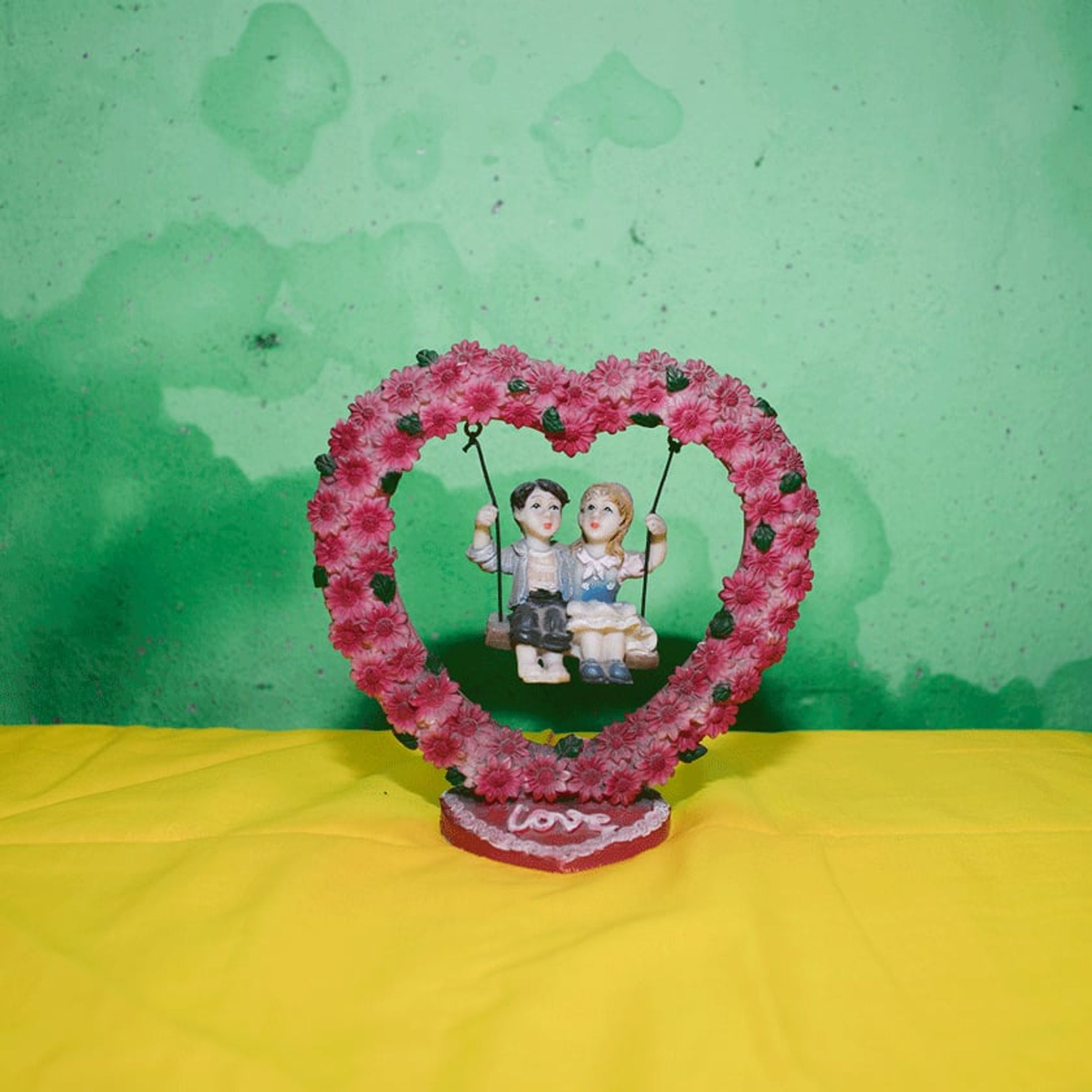 Heart-shaped floral ornament with a miniature couple sits on a bright yellow cloth.