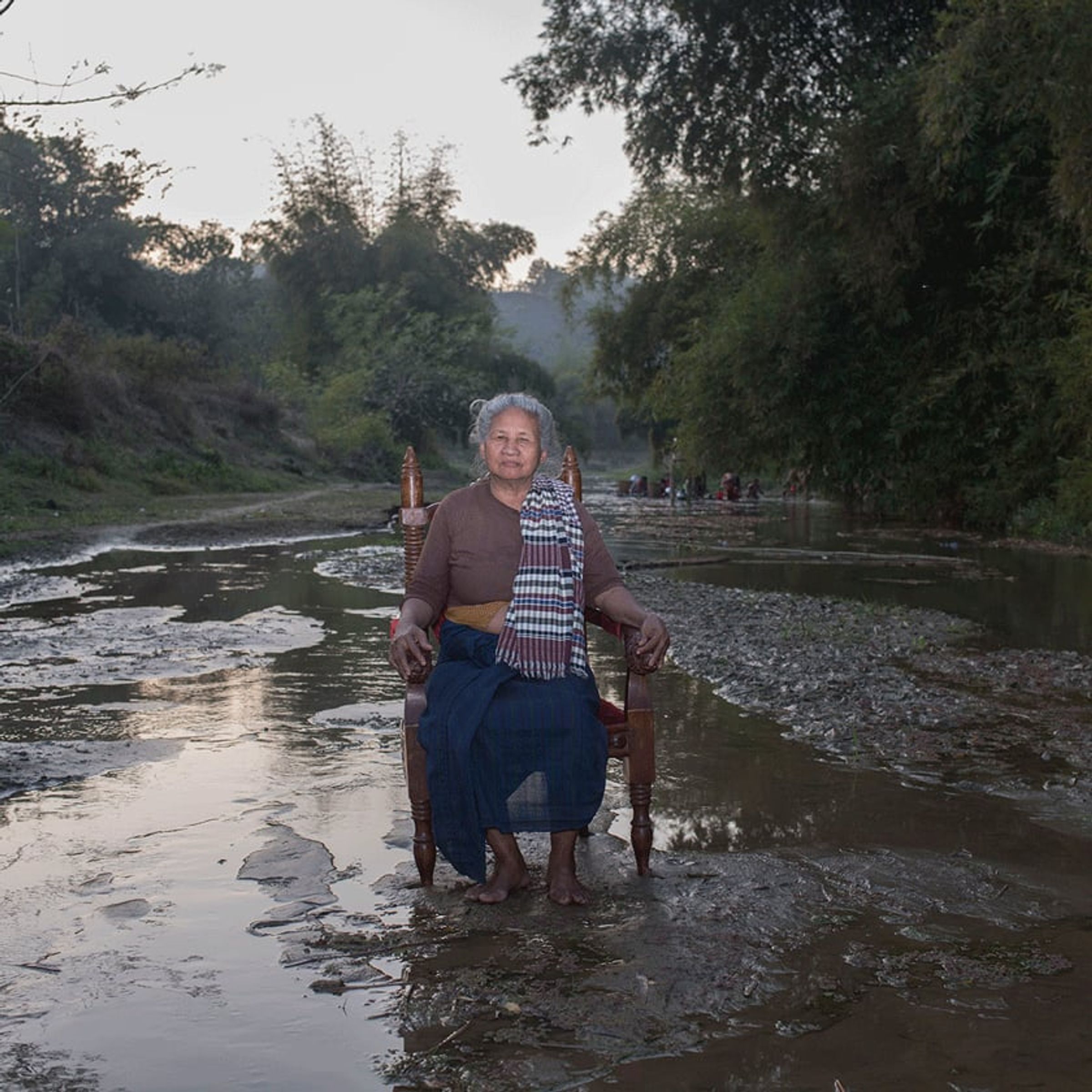 Older person seated beside a wooden chair in a shallow rocky river.