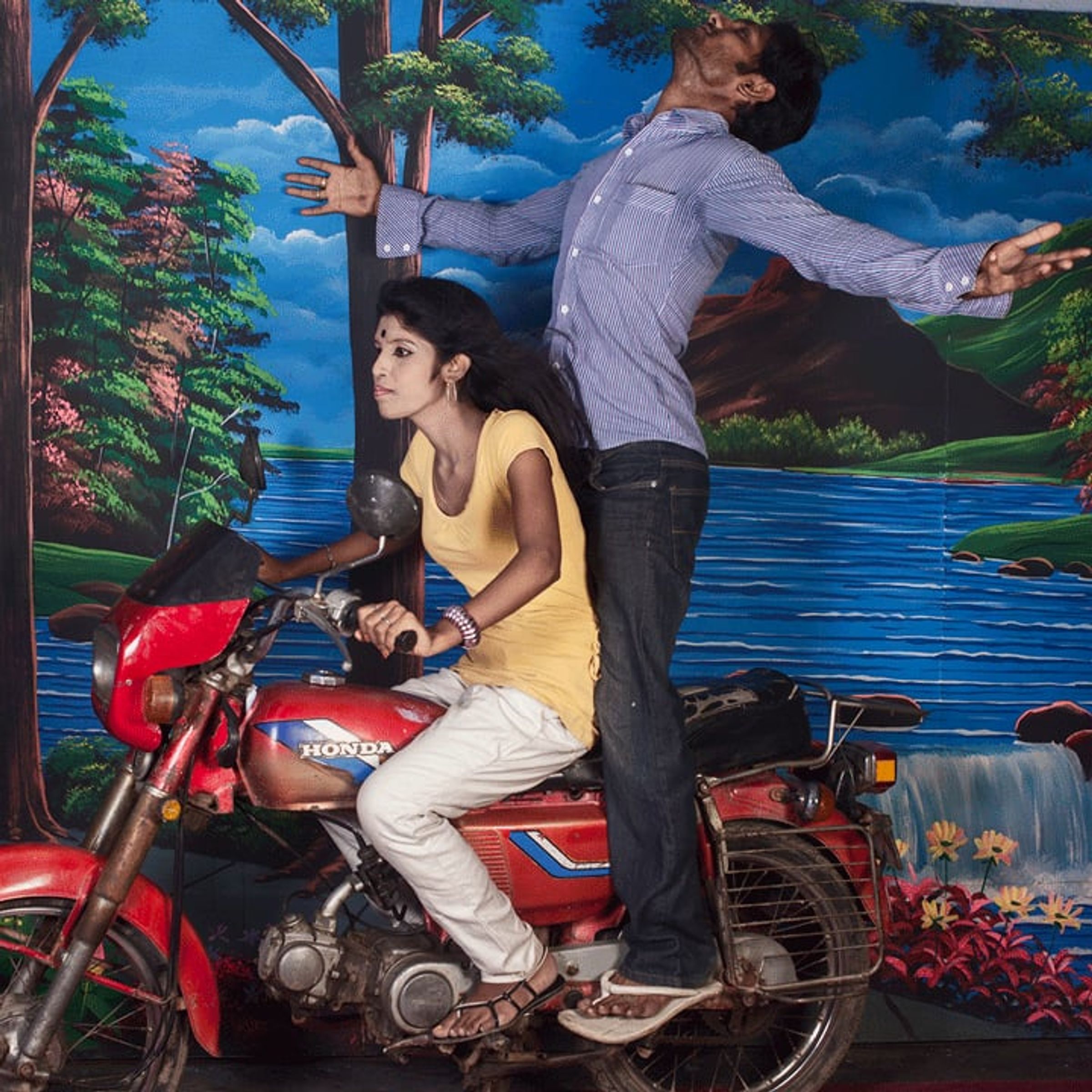 Woman sits on a red motorcycle while a man behind her stretches both arms outward.