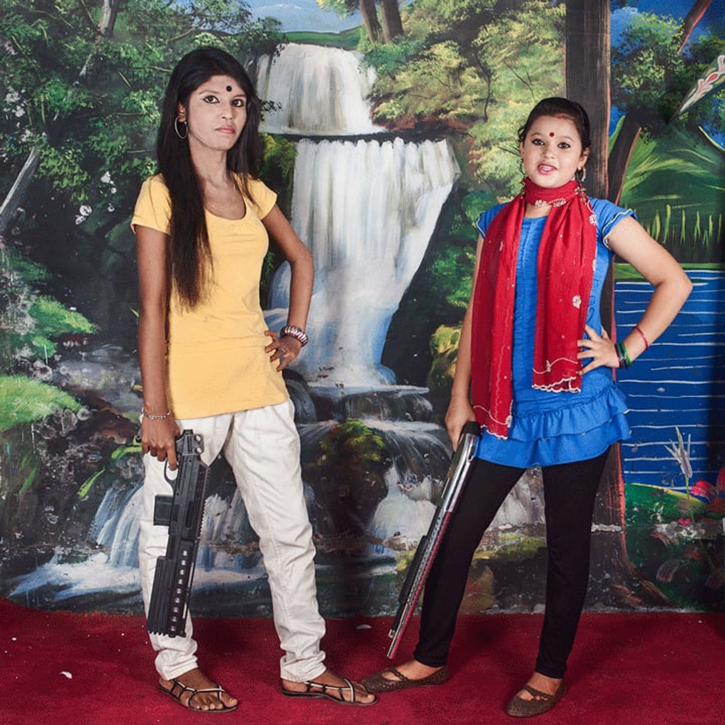 Two young people pose with toy guns before a painted waterfall.