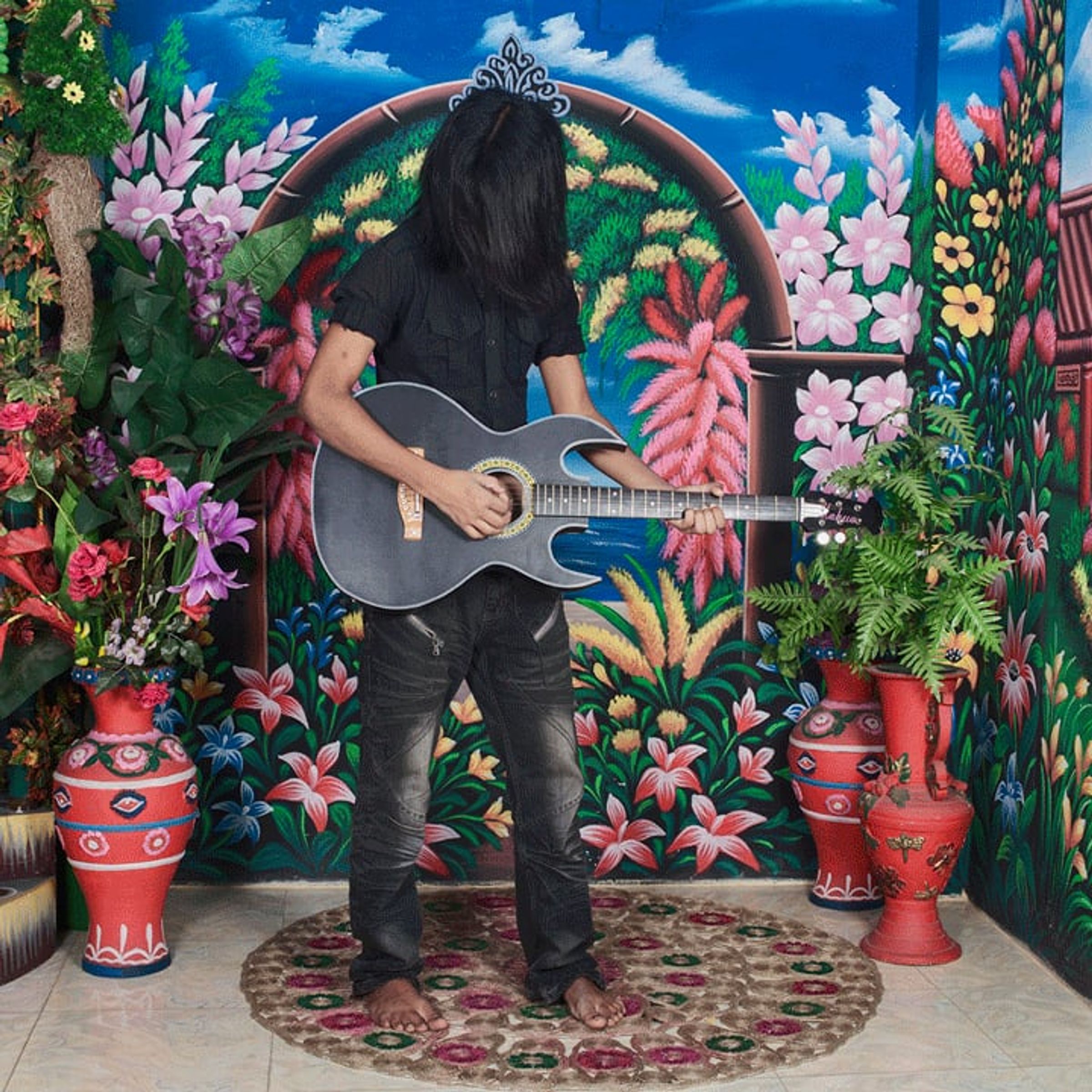 Long-haired guitarist faces away before a brightly painted floral garden backdrop.