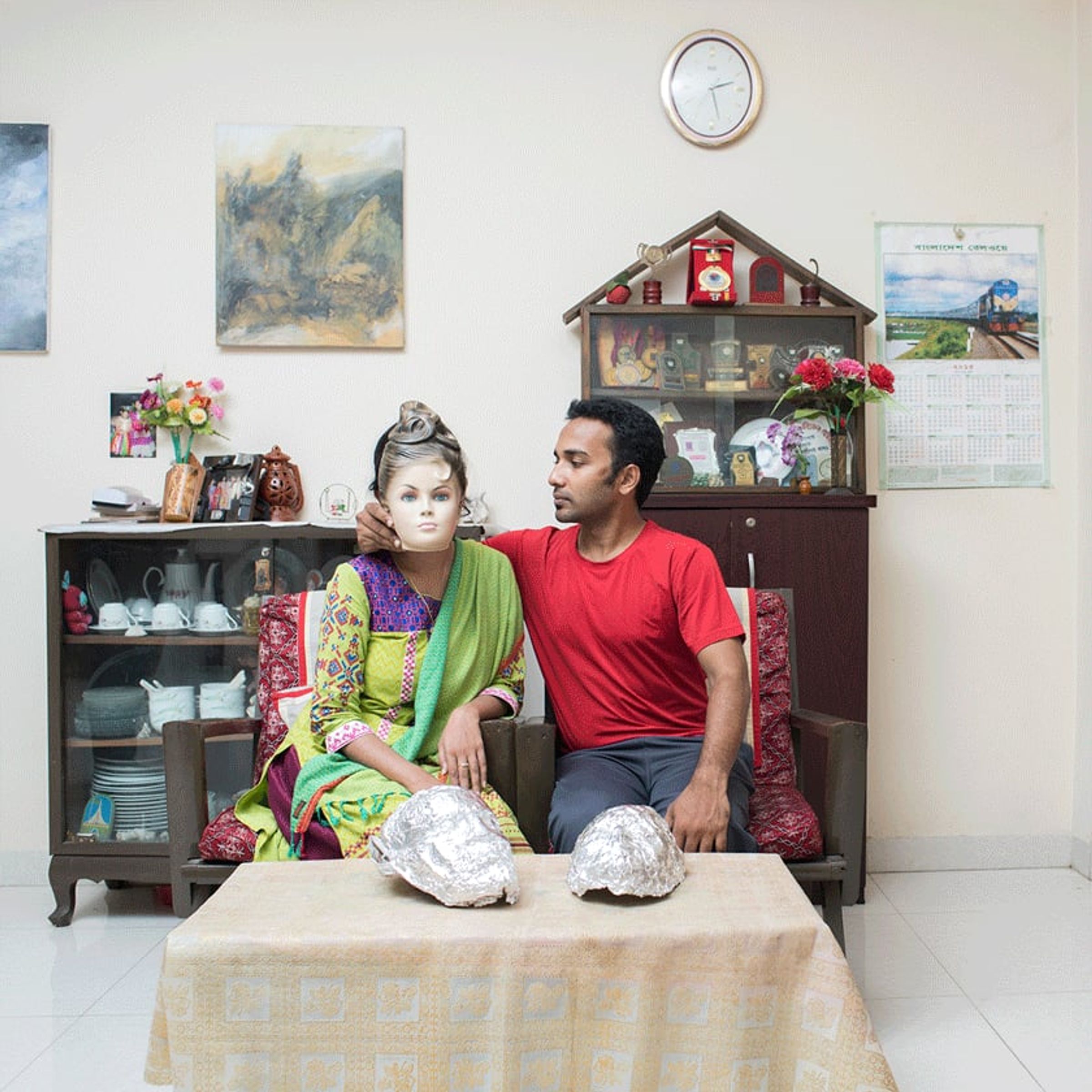 Seated couple in a living room, one wearing a mannequin-like mask beside two foil-wrapped objects.