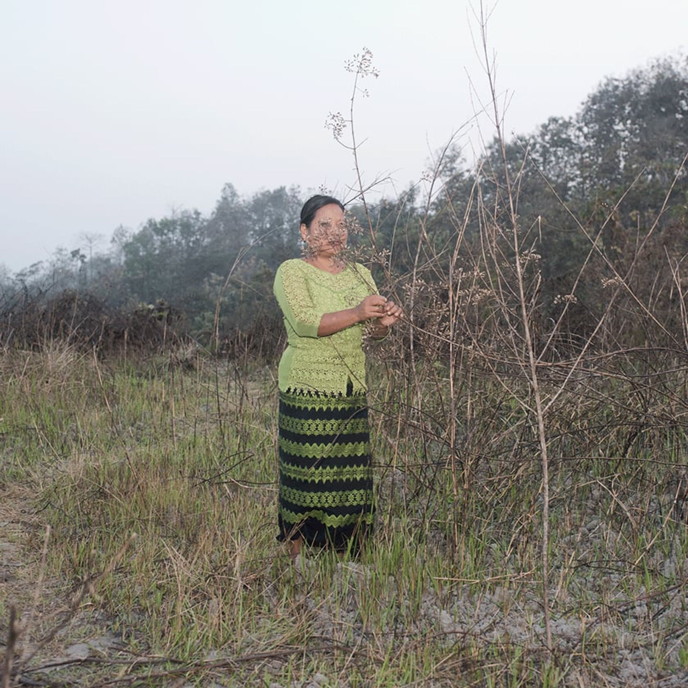 Person in green patterned dress holds brittle branches in a dry field.