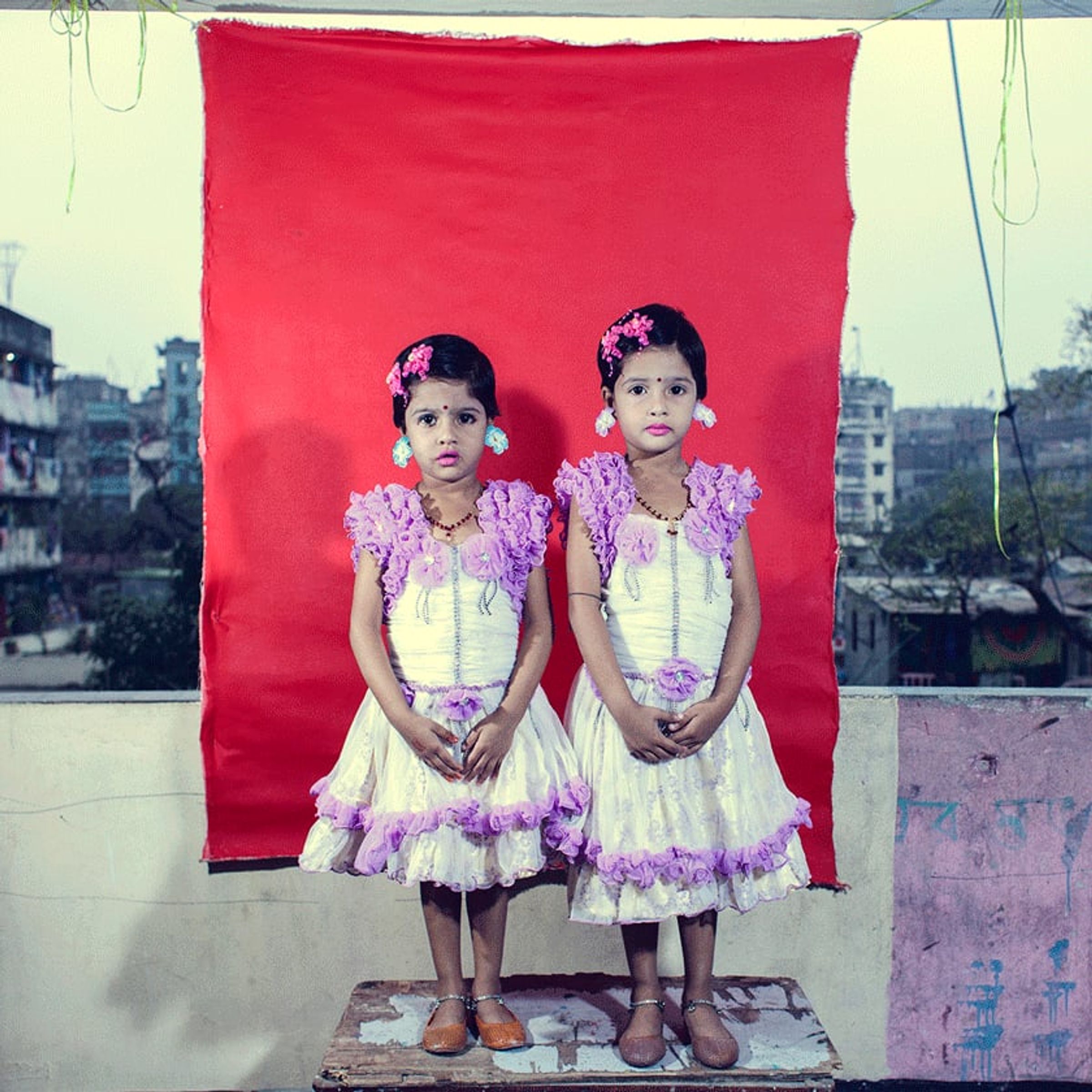Two children in matching pink dresses stand side by side before red cloth.