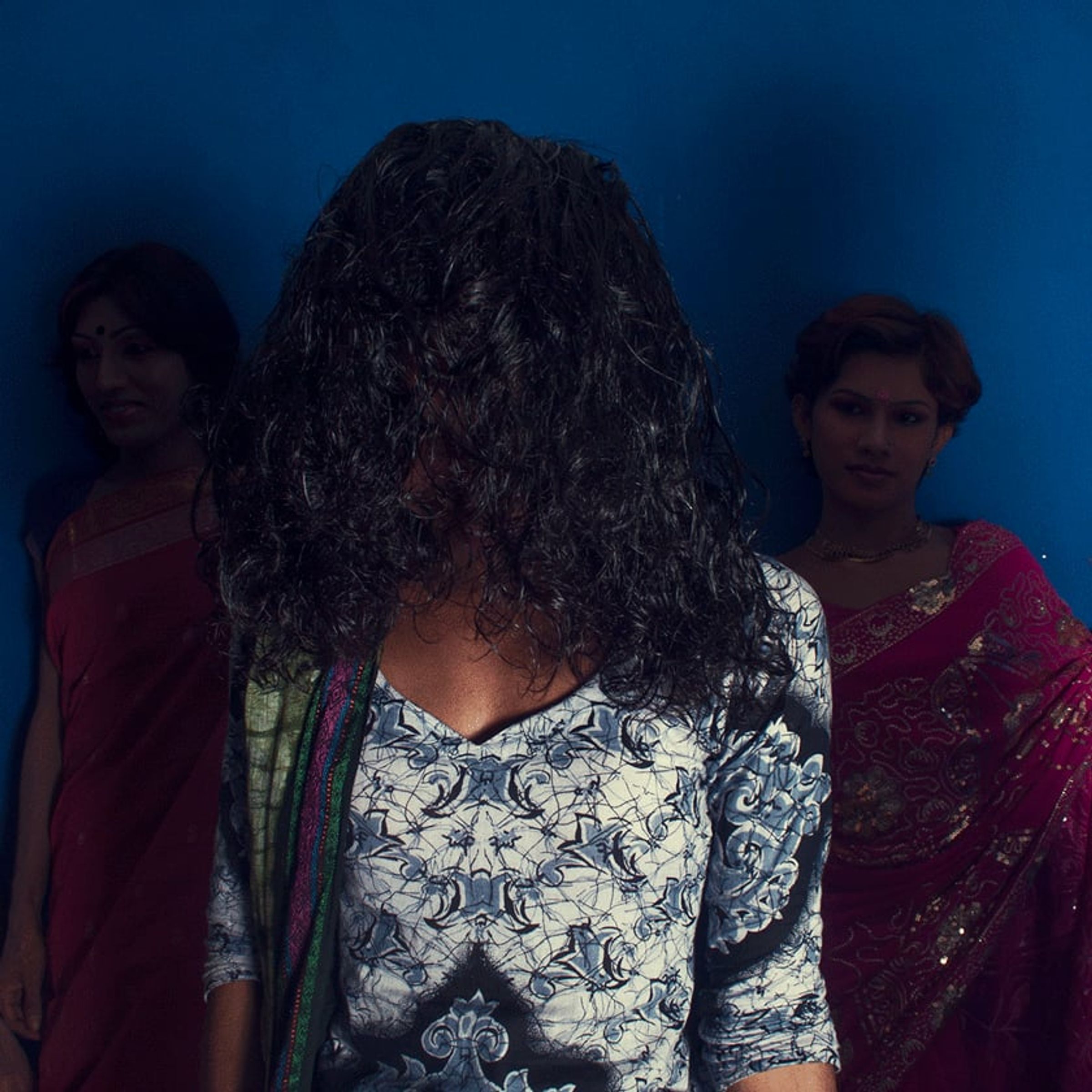 Person with long black hair faces away while two brightly dressed figures stand against blue.