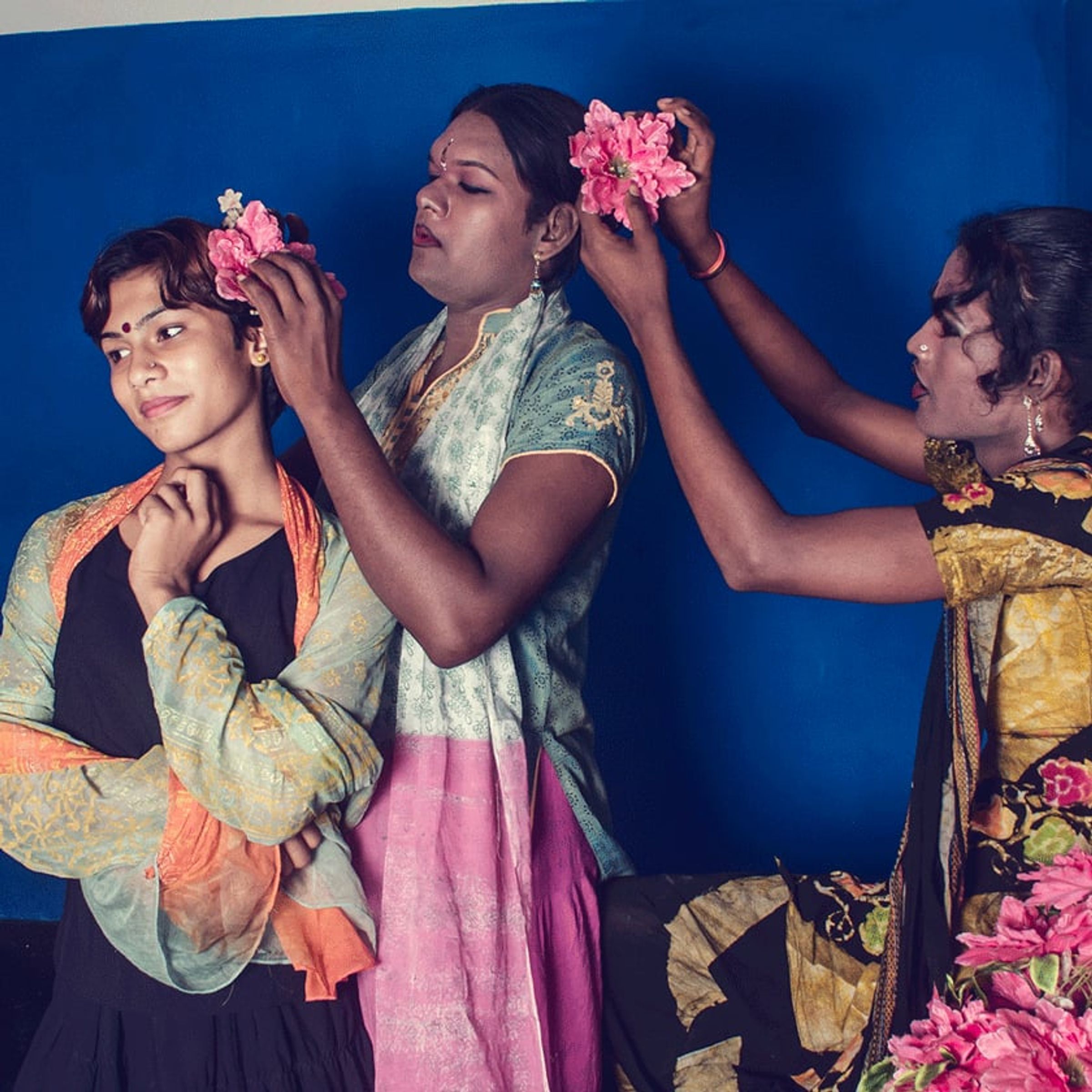 Three people arrange pink flowers in the hair of a seated person.