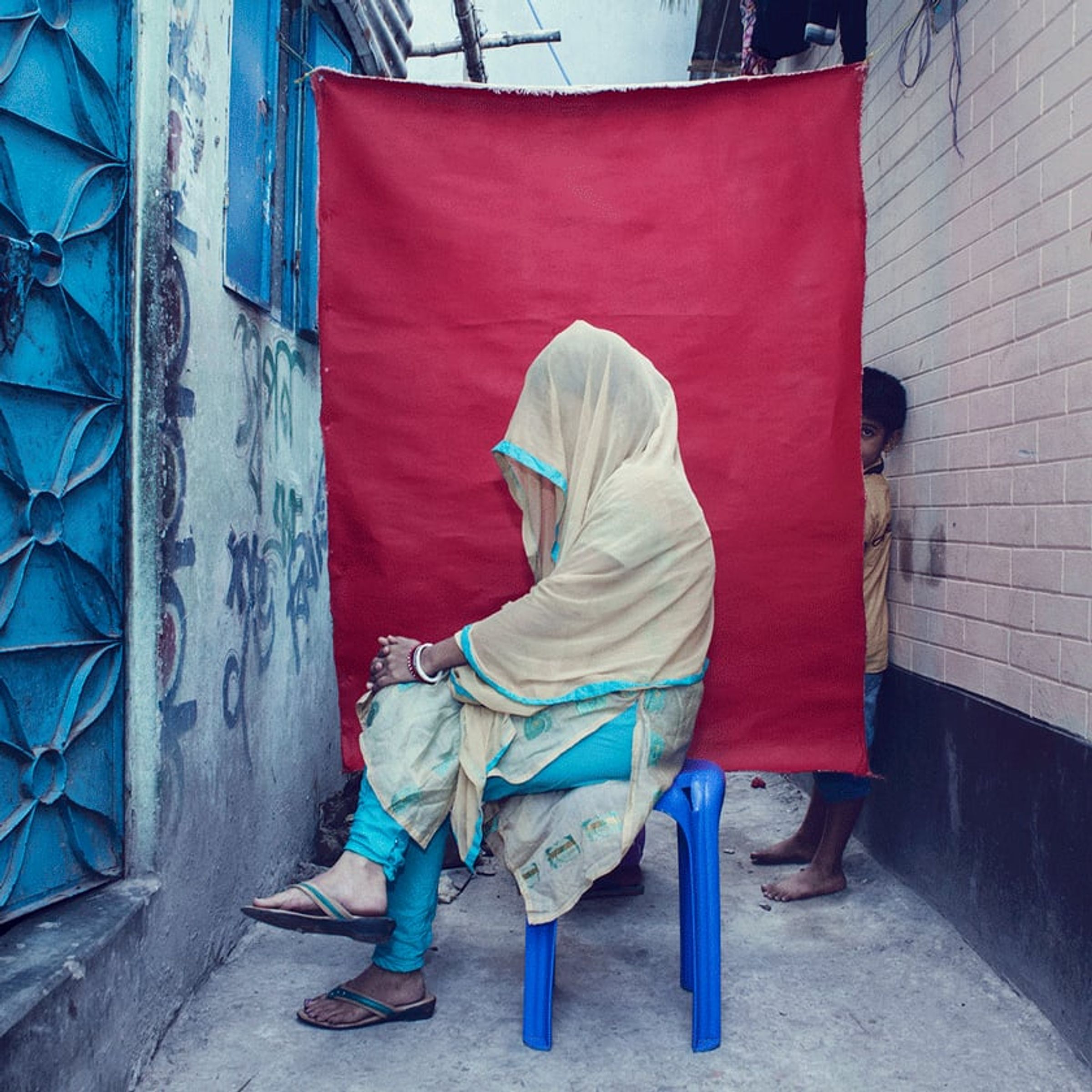 Veiled person sits sideways on a blue stool against red fabric in a narrow alley.