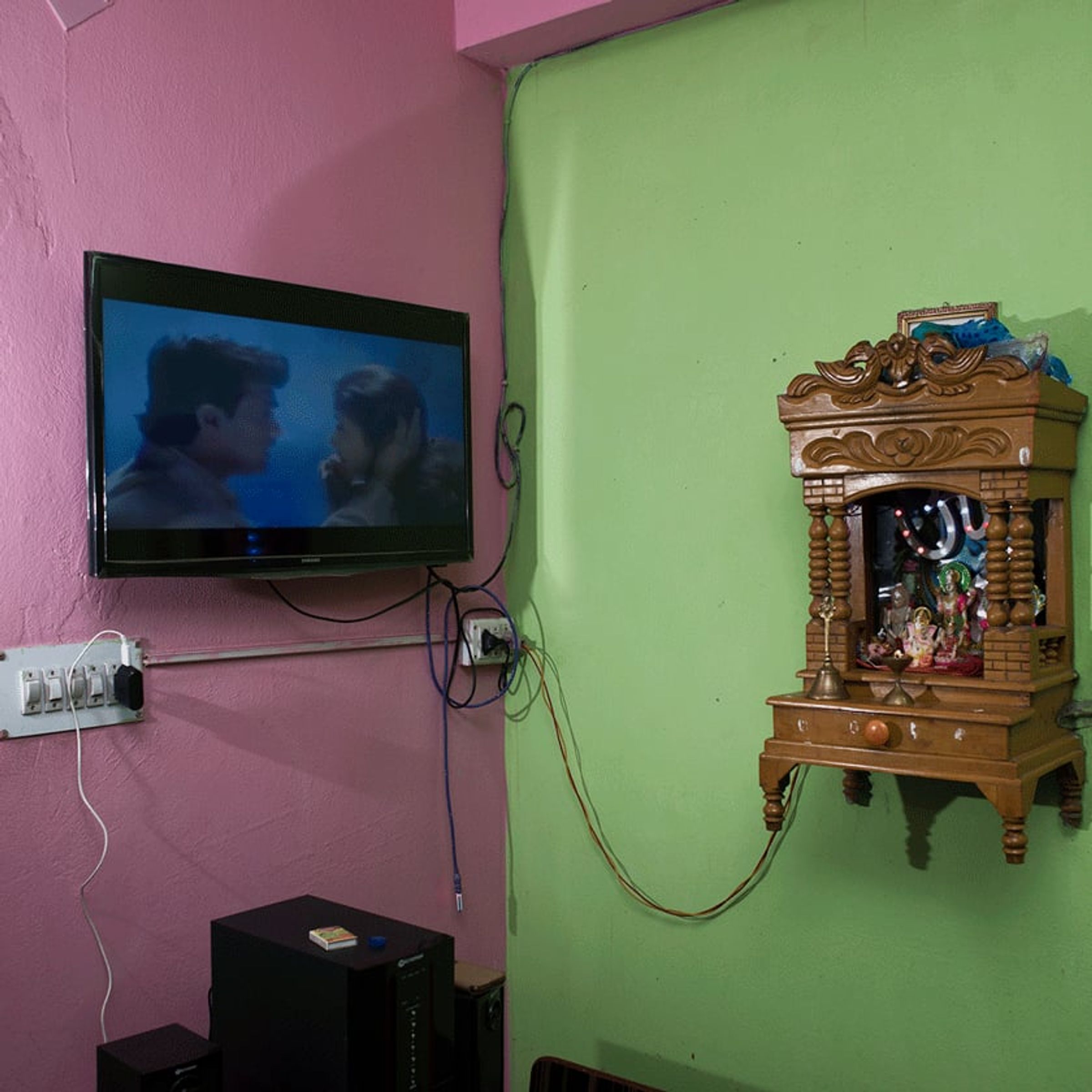 Television and a small wooden shrine are mounted on adjoining pink and green walls.