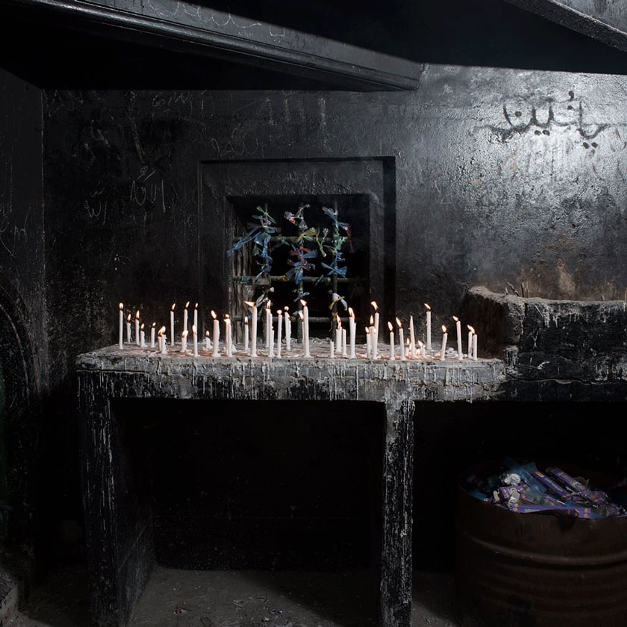 Burning candles line a soot-blackened shelf beneath hanging ornaments and wall script.