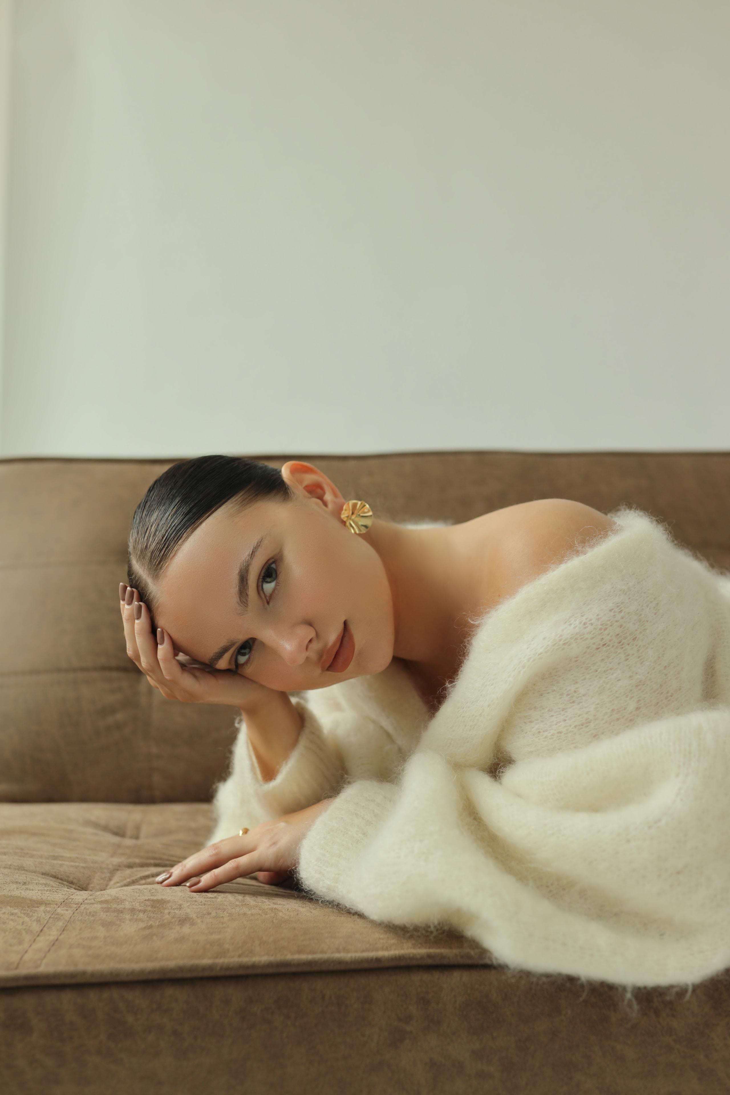 Model resting head on hand on tan sofa in white off-shoulder mohair sweater, soft natural light