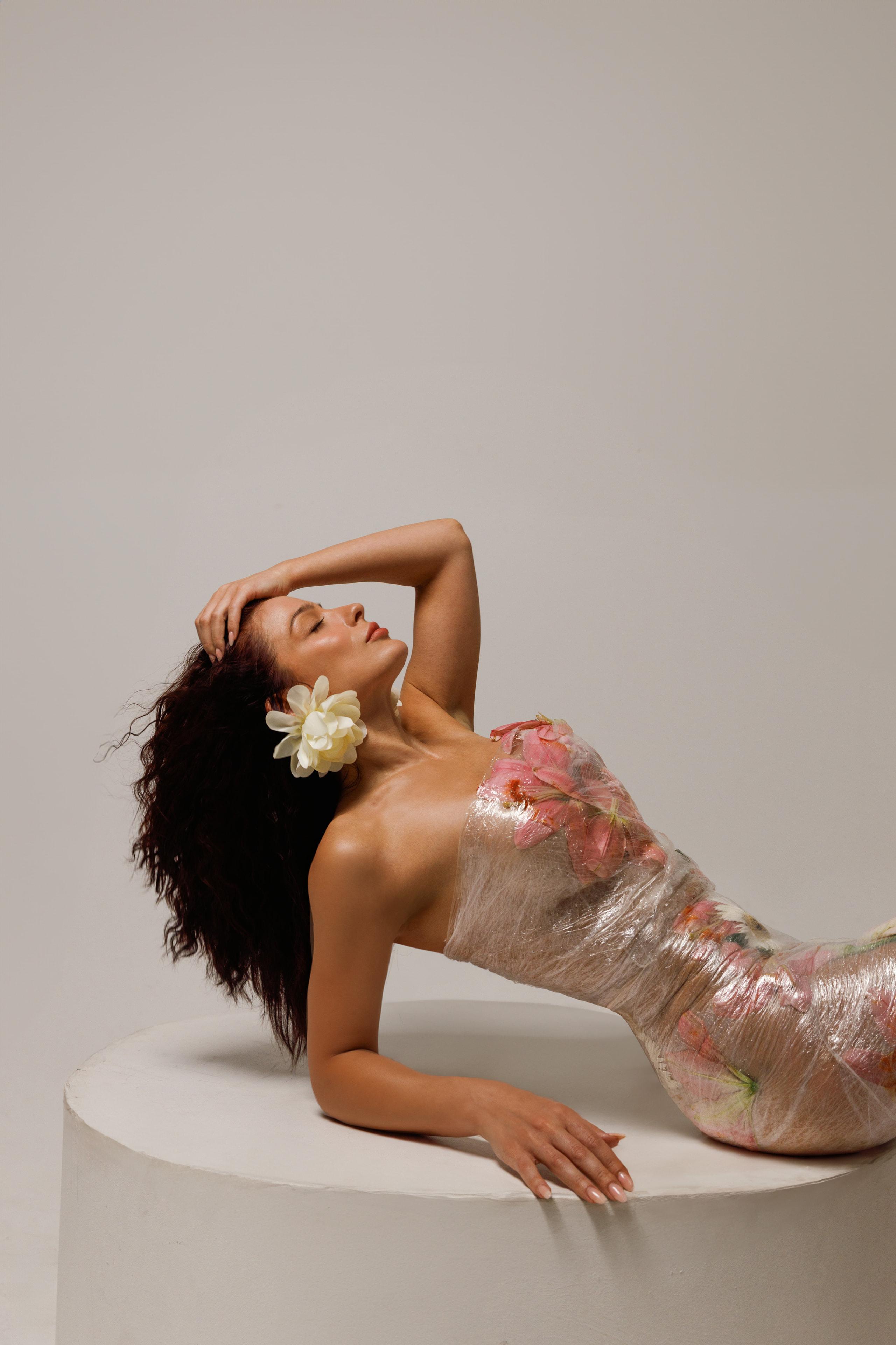 Brunette woman reclining on white plinth wrapped in floral cellophane dress with flower behind ear