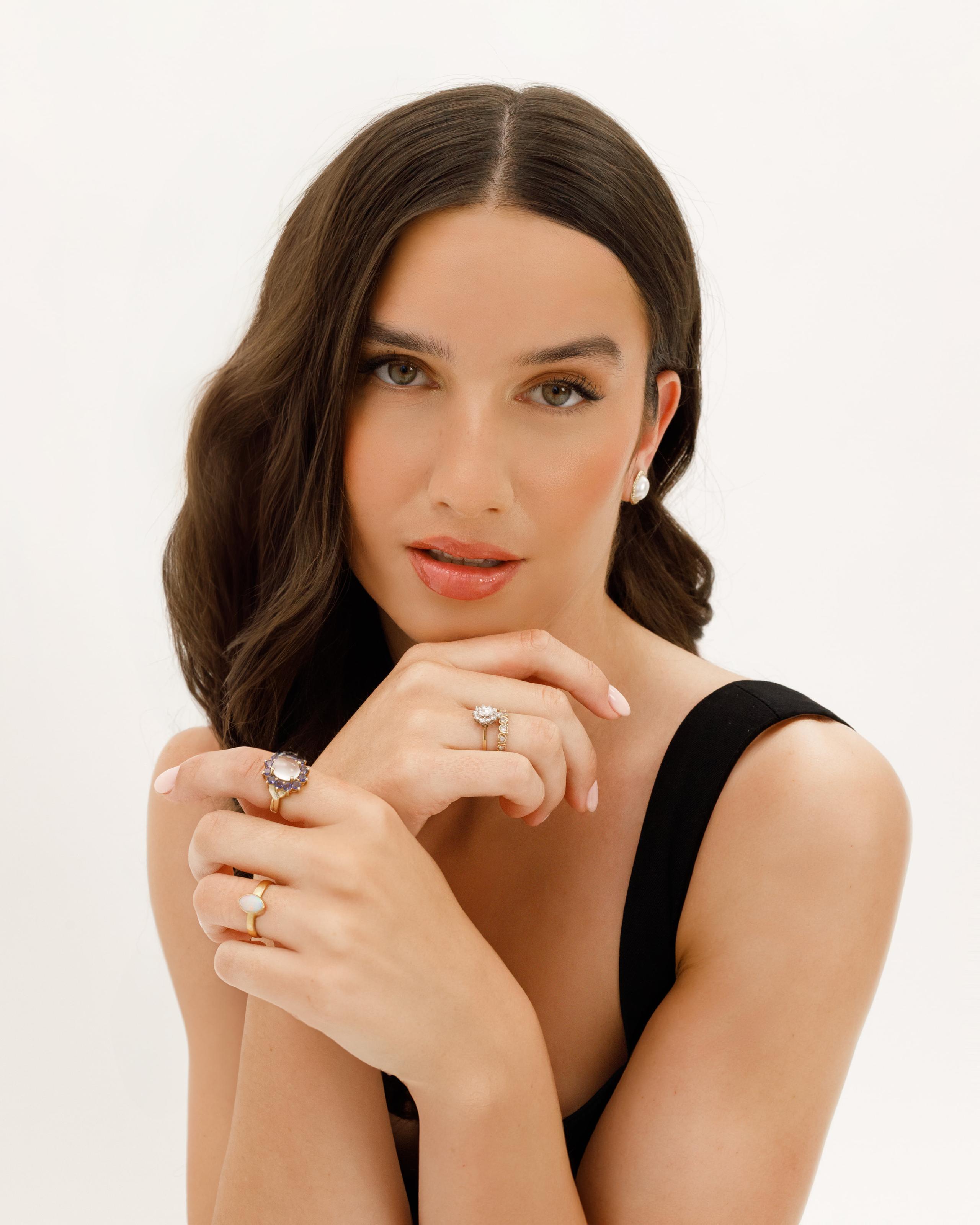 Woman in black top resting chin on hands, wearing pearl earring and several gemstone and gold rings