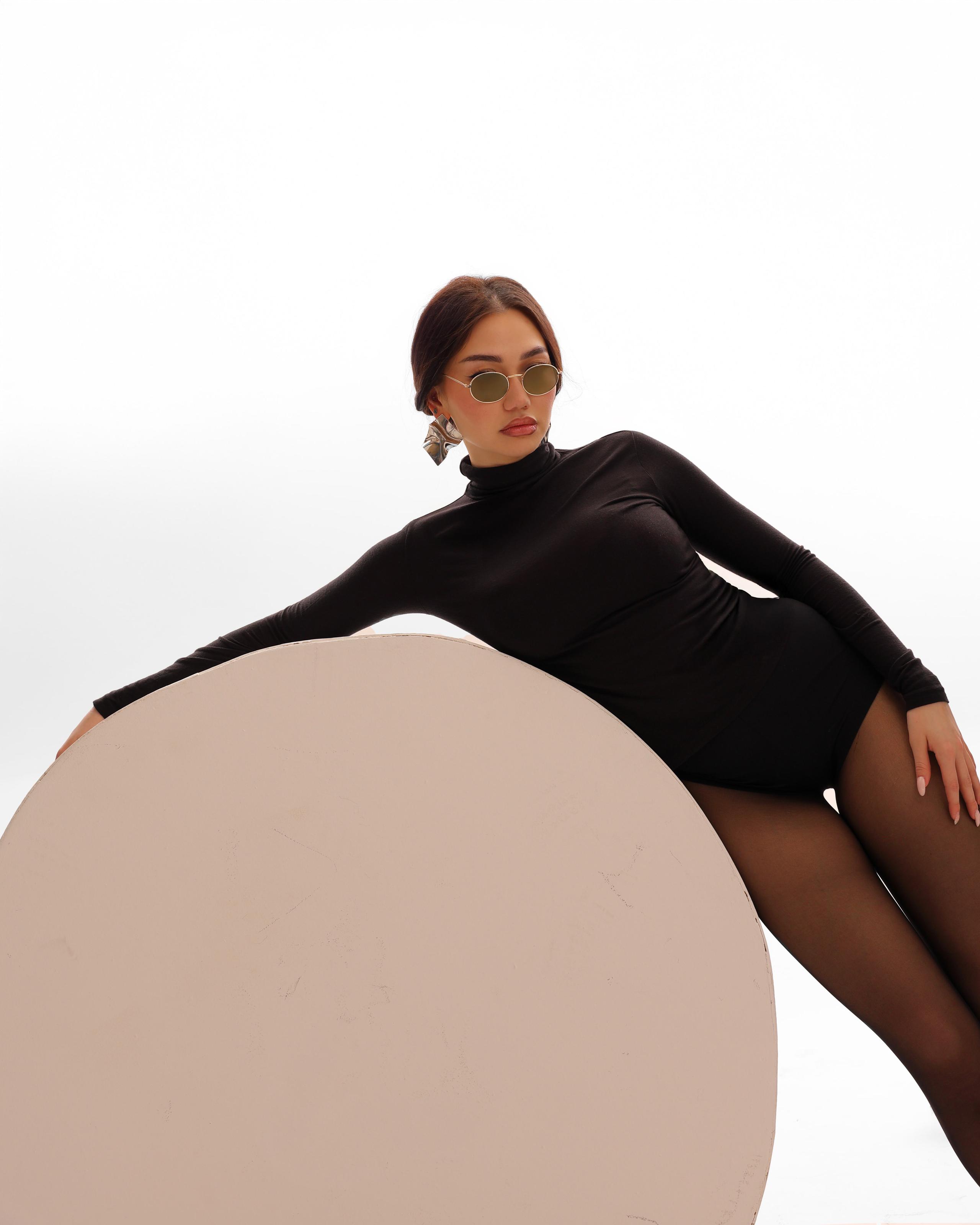 Model in a black bodysuit and tinted sunglasses reclines against a large round beige prop on white
