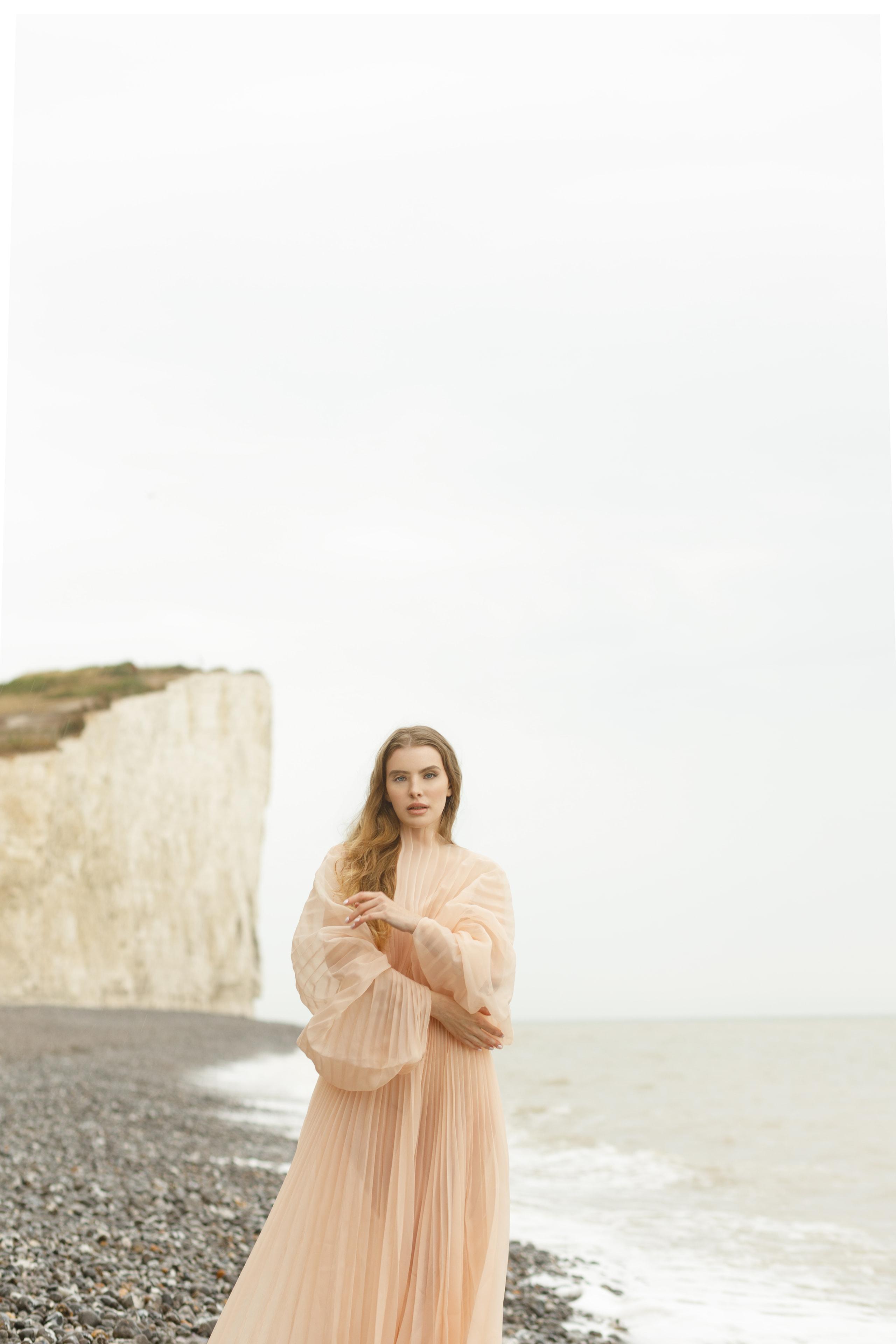 Woman in flowing peach pleated gown standing on pebble beach below chalk cliffs, soft overcast light