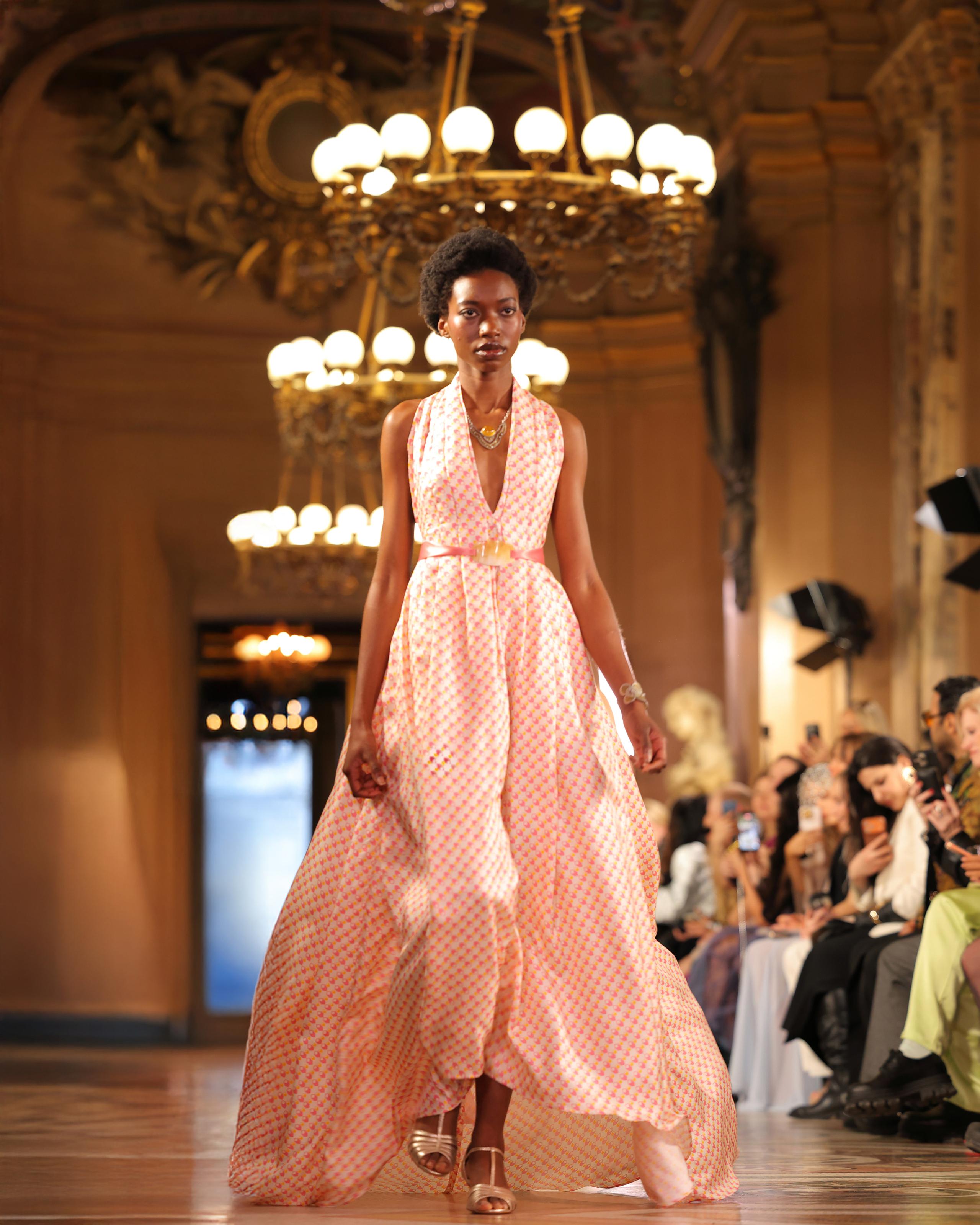 Model in flowing blush patterned halter gown beneath chandeliers on runway